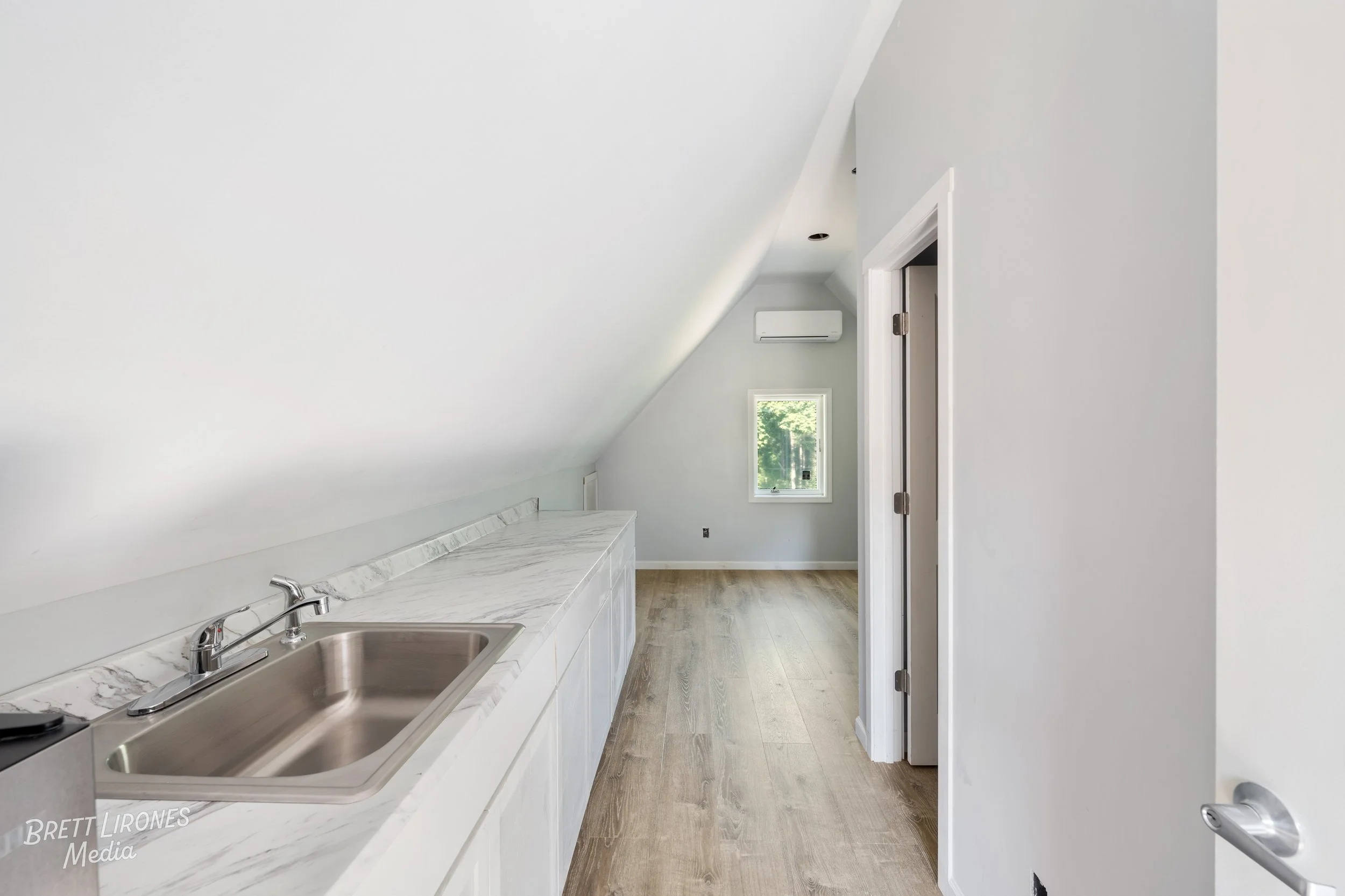 Empty kitchen with white cabinets, marble countertops, stainless steel sink, wood flooring, small window, and an air conditioning unit on the wall.
