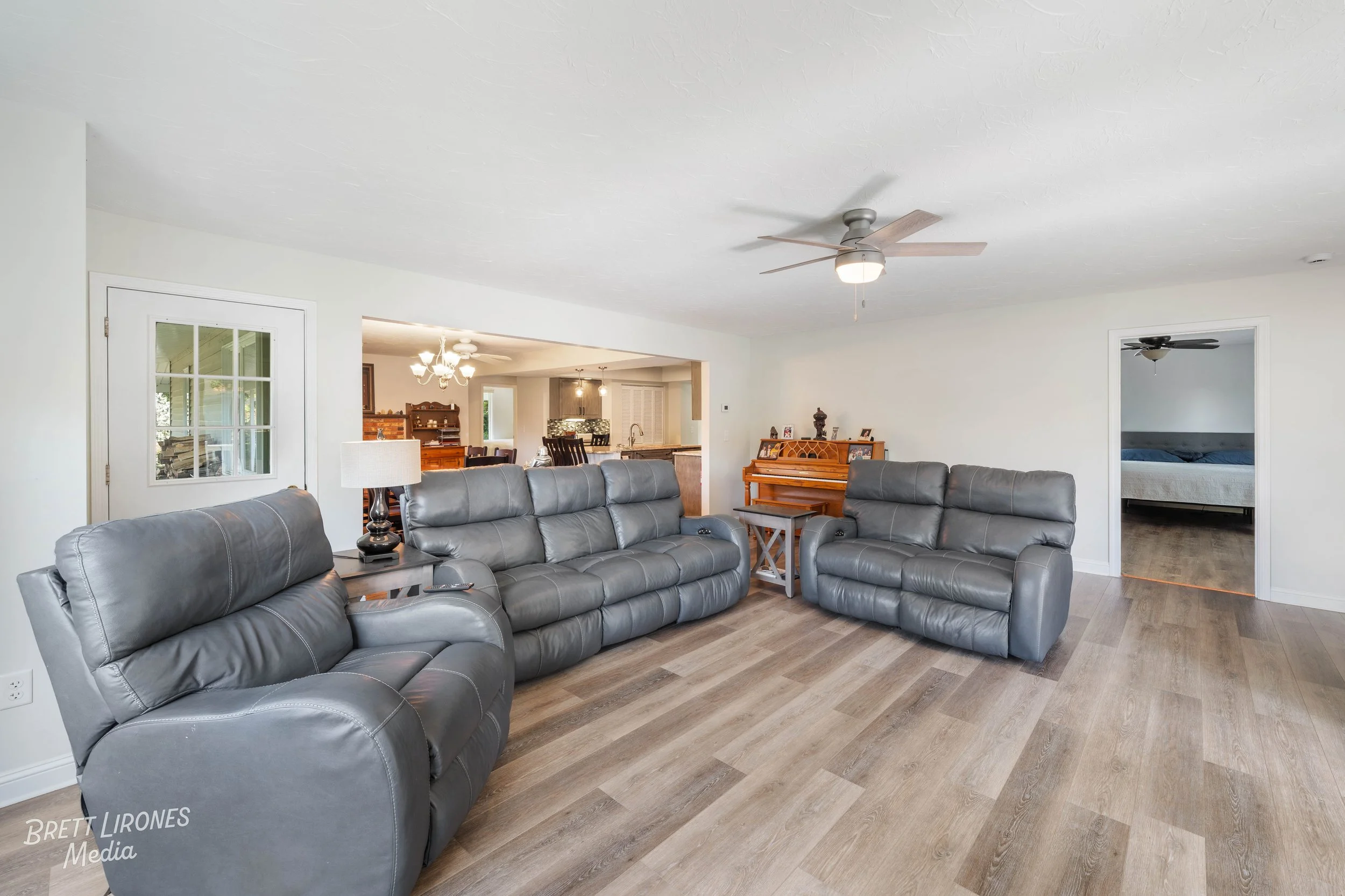 Living room with three gray leather recliner sofas, a side table with a lamp, a wooden piano against the wall, and a view into the dining area and a bedroom.