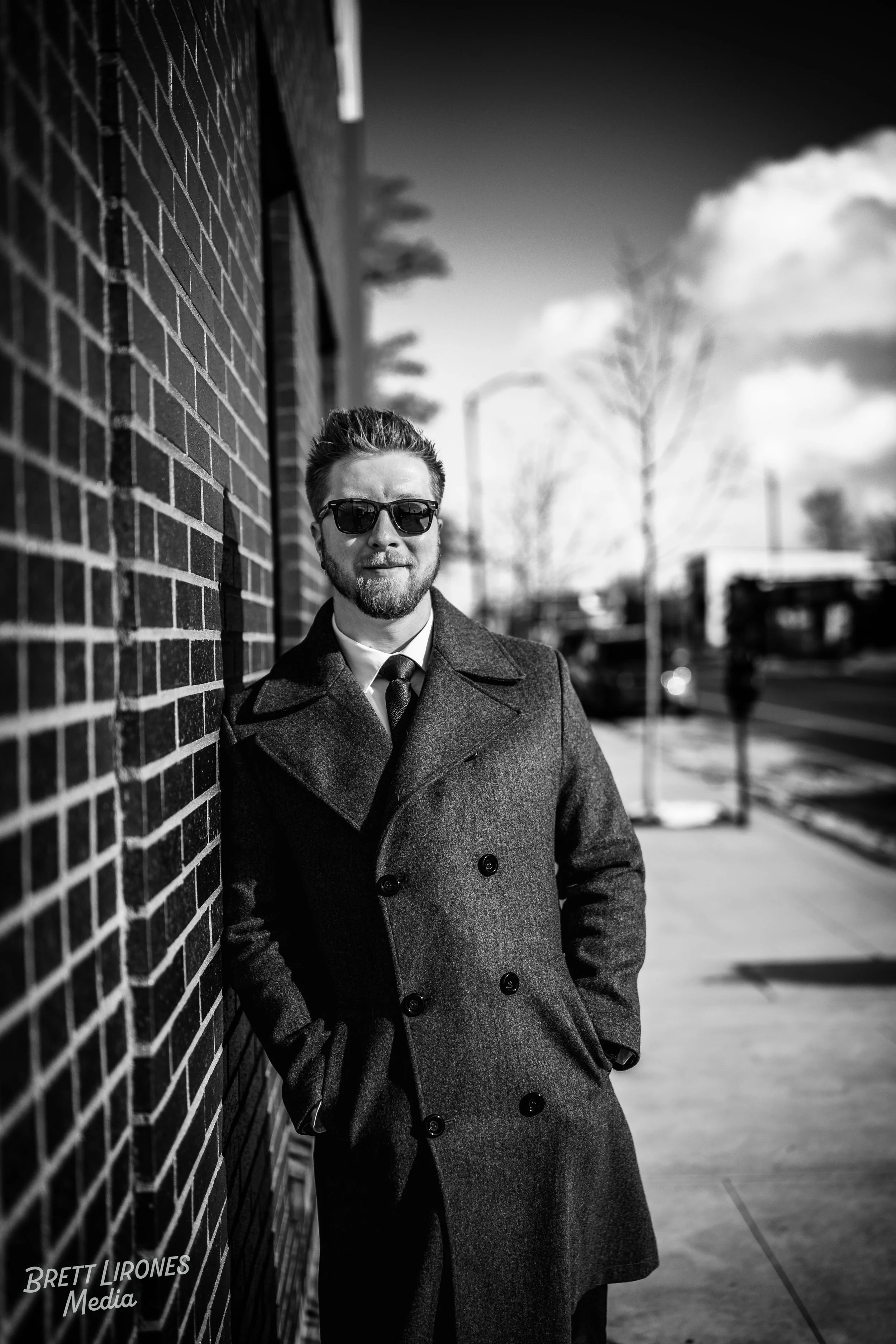 A man with glasses and a beard leaning against a brick wall on a city sidewalk, wearing a dark overcoat and tie, with a blurred background of trees and people, in black and white.