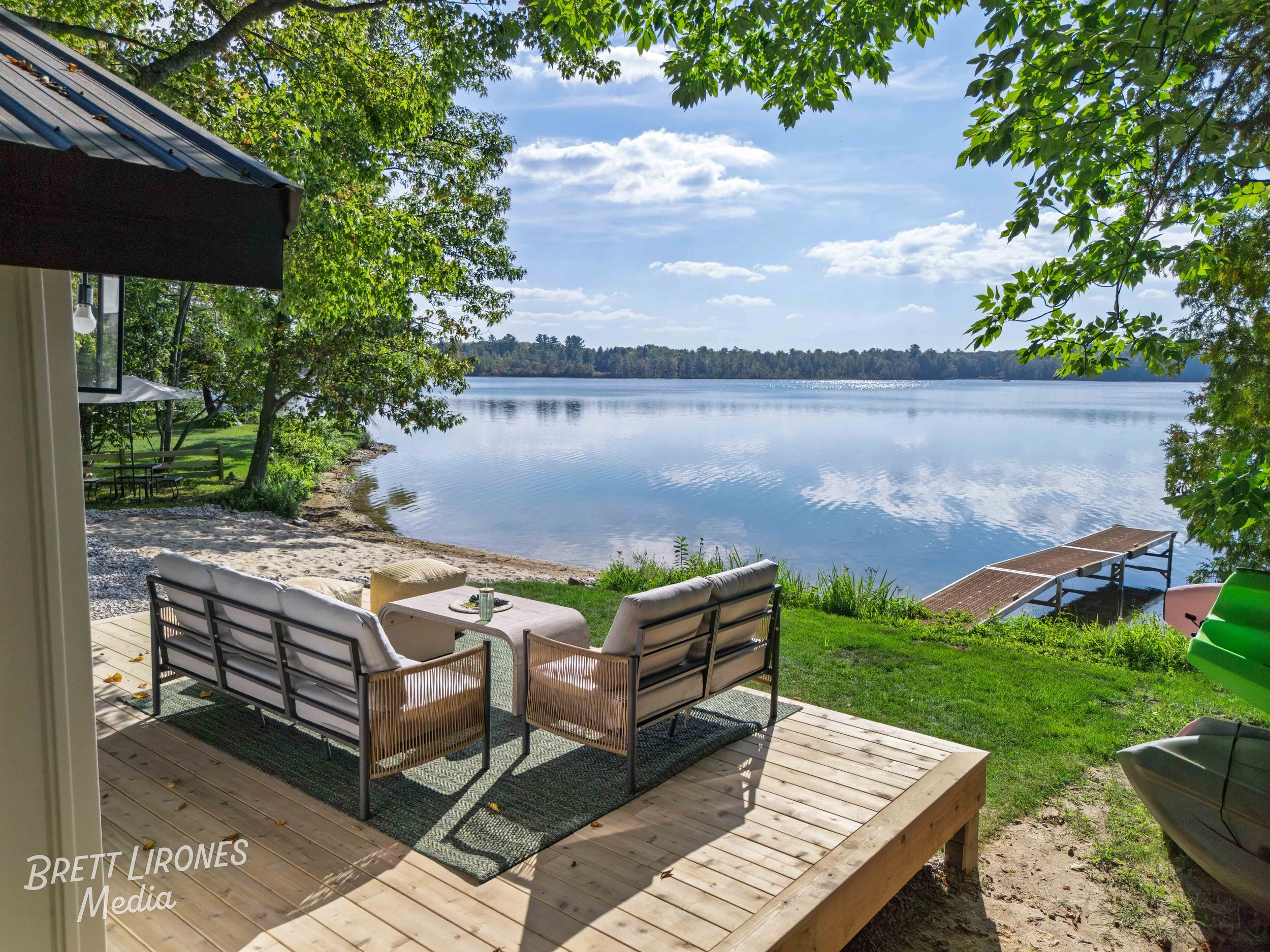 View of a lakeside patio with outdoor seating on a wooden deck, overlooking a calm lake, surrounded by green trees, with a dock extending into the water and a blue sky with scattered clouds.