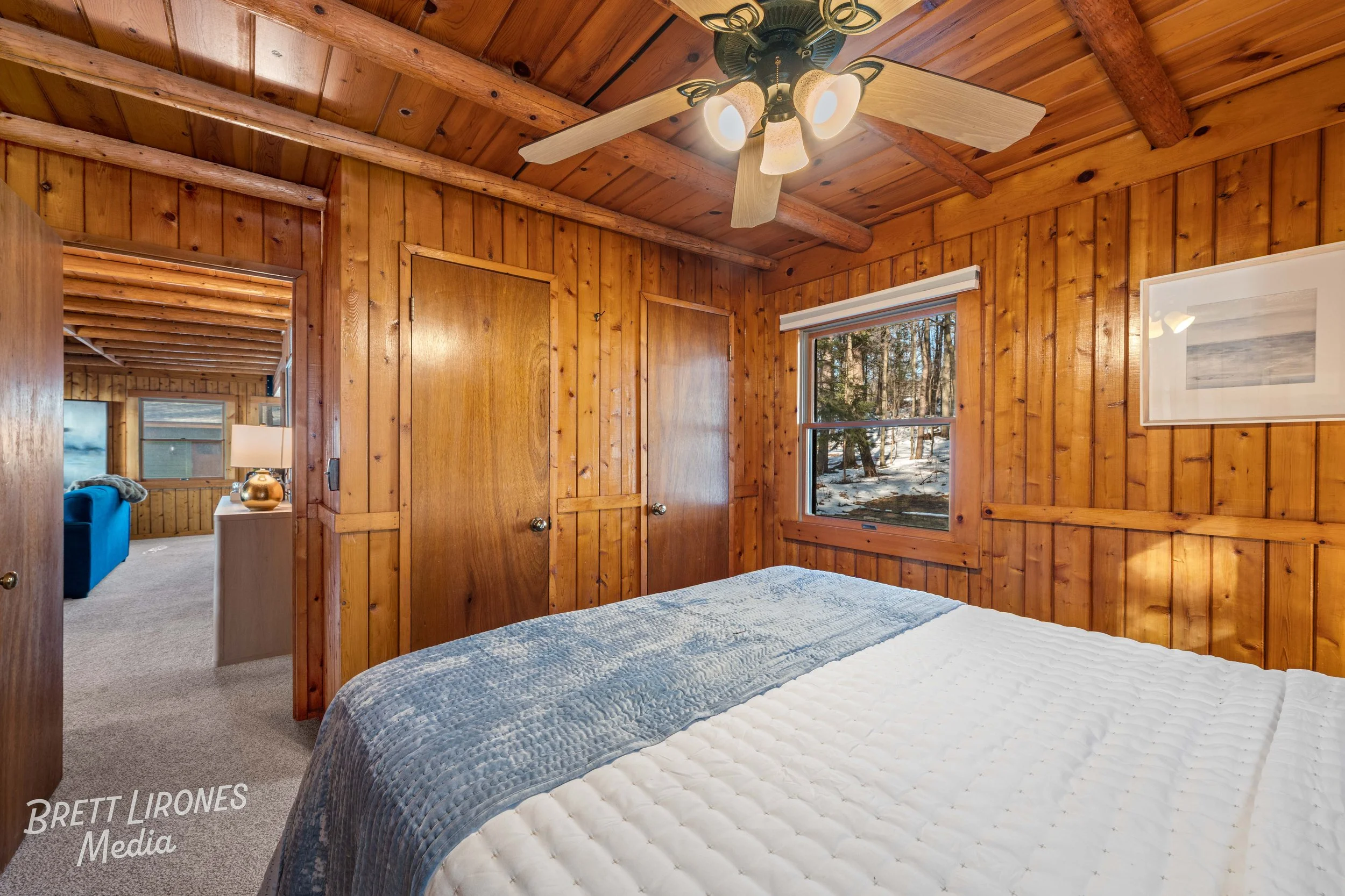 A cozy bedroom with wooden paneled walls and ceiling, a window showing a wooded outdoor scene, a ceiling fan with lights, a bed with a white quilt and a blue blanket, and a framed picture on the wall.