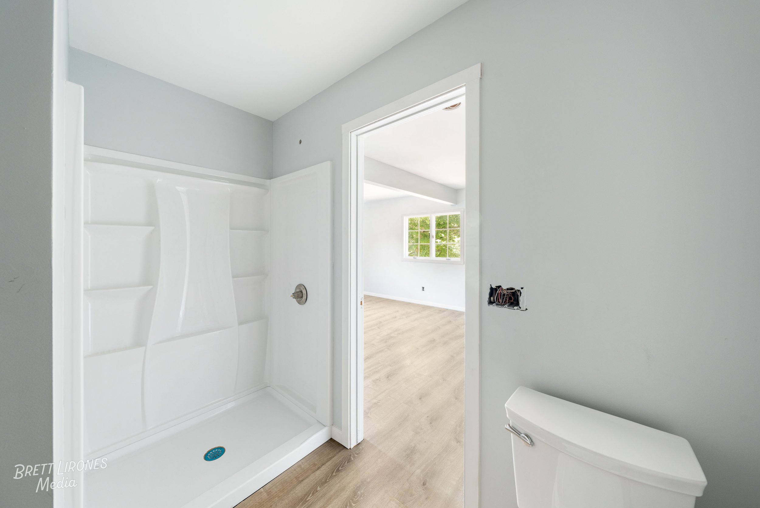 Empty bathroom with a white shower stall and a toilet, with an opening leading to a bright room with windows.