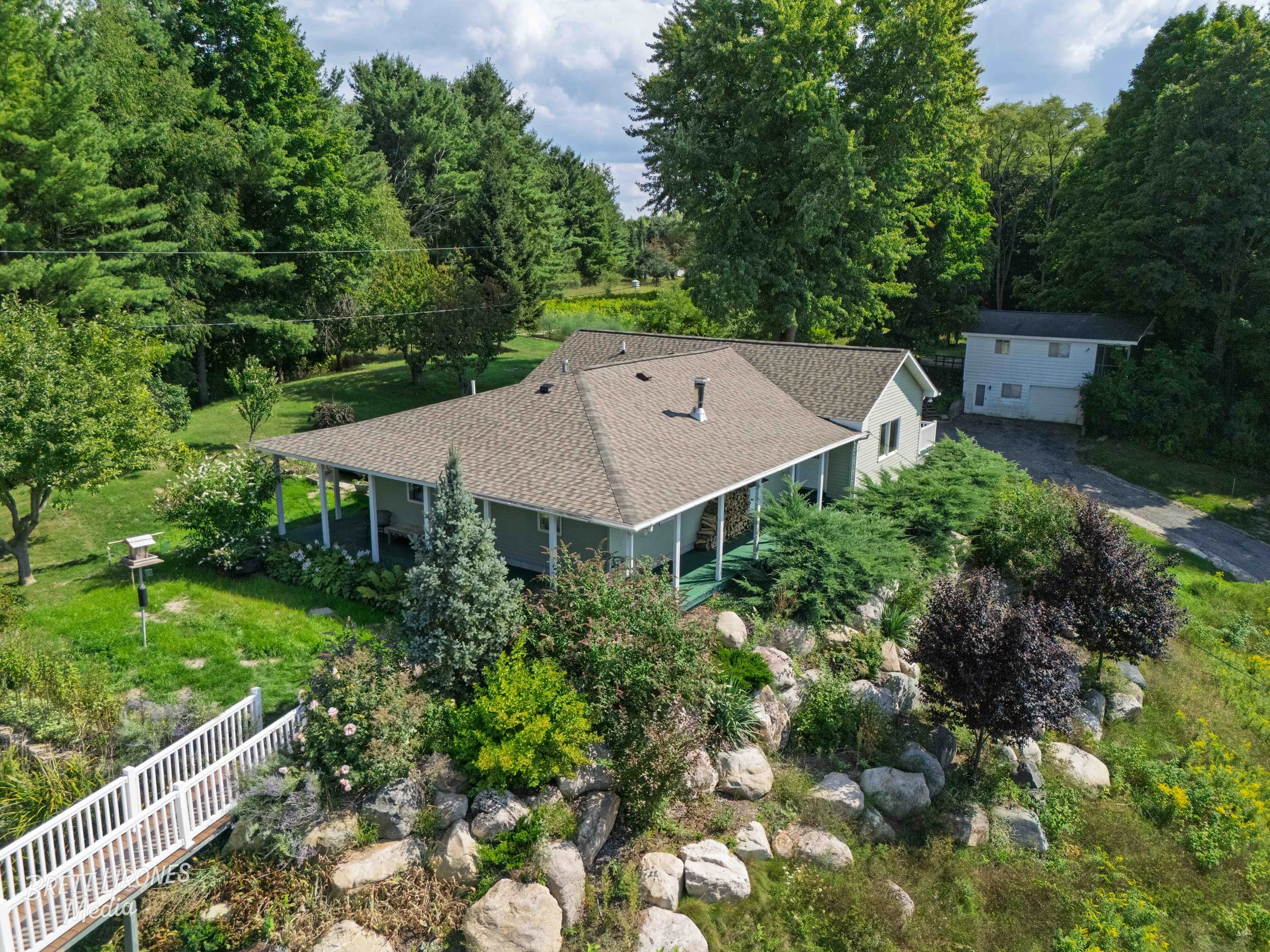 A house with a brown roof and gray walls surrounded by lush green trees and landscaped garden, with large rocks lining the garden border and a white fence in the foreground.