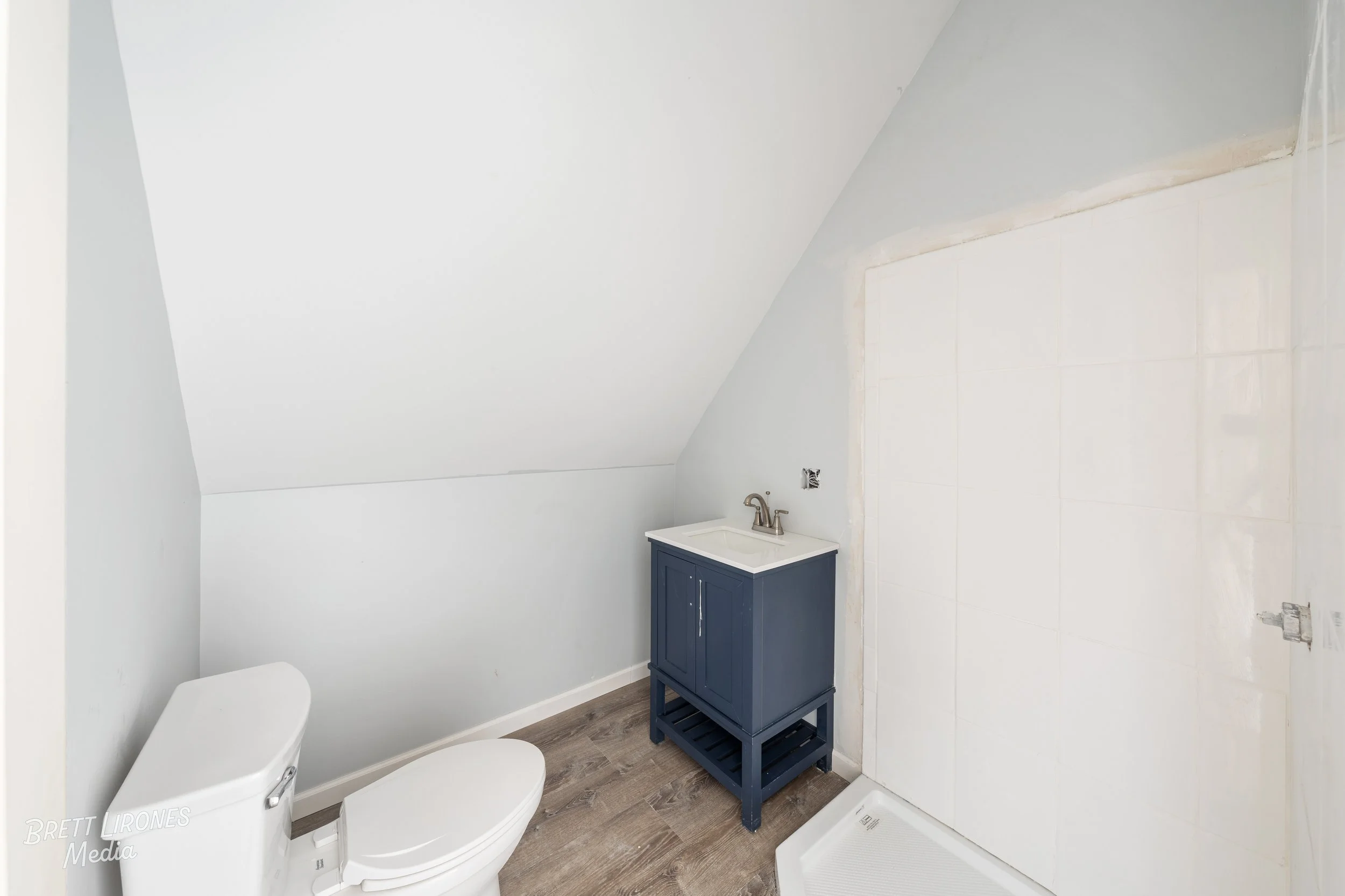 Small bathroom with white walls, a toilet, a blue vanity with a small sink, and a shower area with white tile walls and a base.