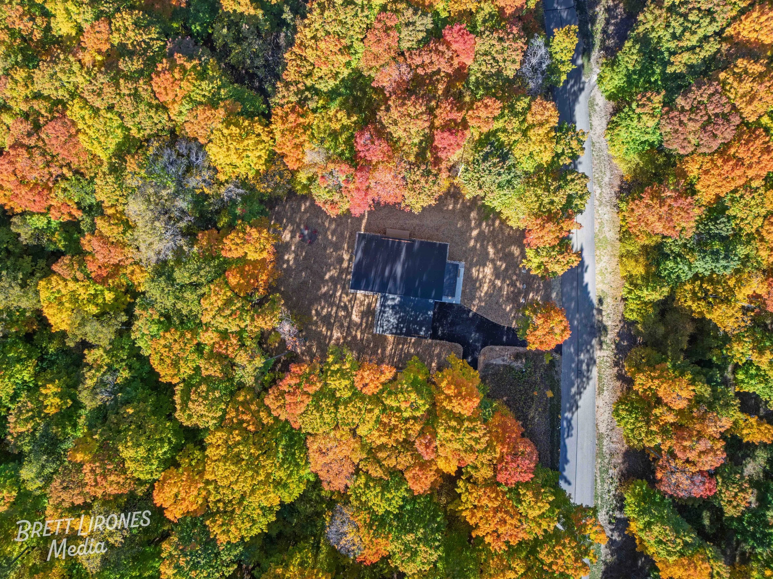 An aerial view of a small building surrounded by vibrant fall foliage in various shades of orange, yellow, red, and green. A dirt driveway encircles the building and leads to a main road on the right side of the image.