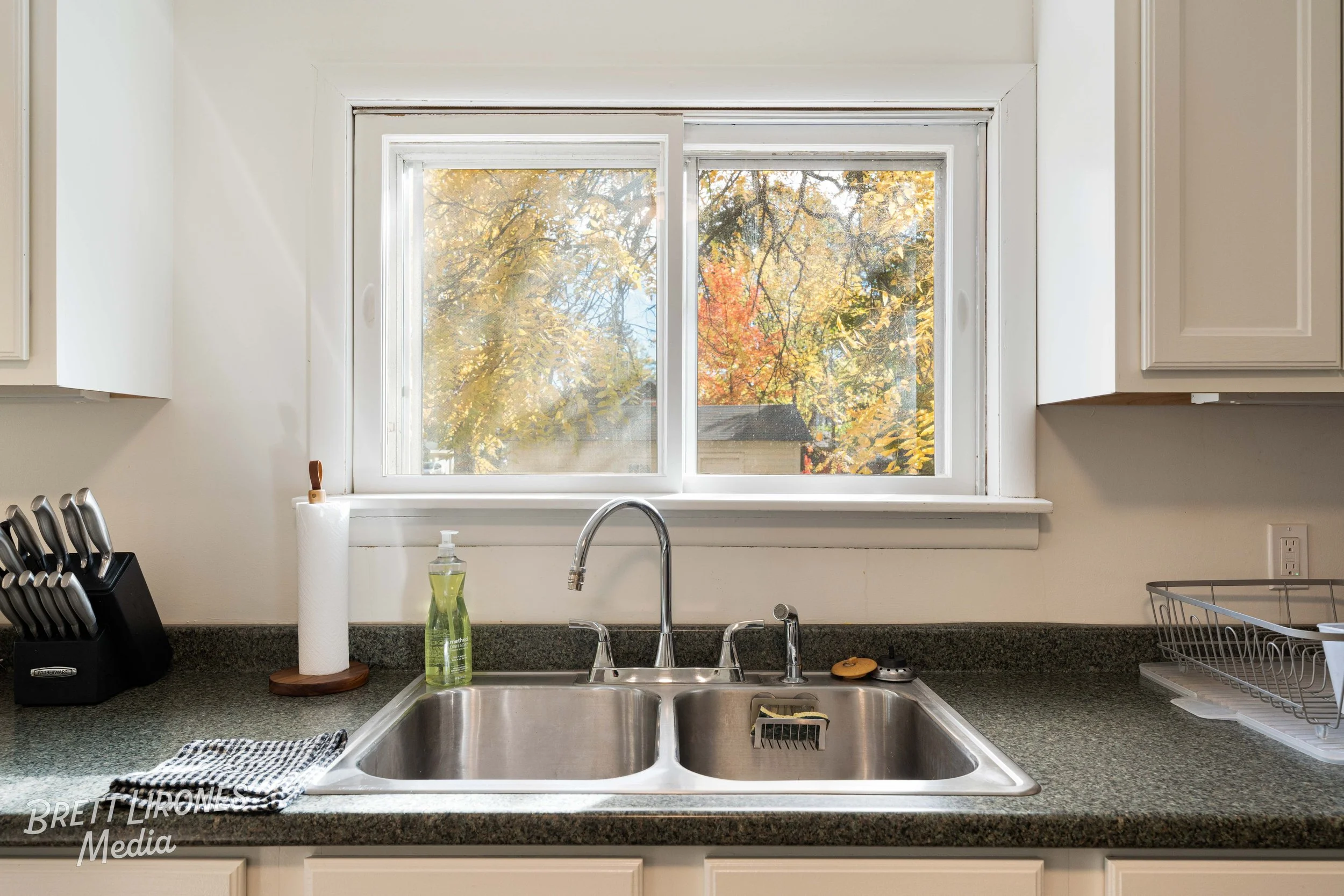 Kitchen sink with two basins and a chrome faucet, a window showing autumn trees outside, a black knife block, paper towel roll, dish soap, dish sponge, and a wire dish rack on the countertop.