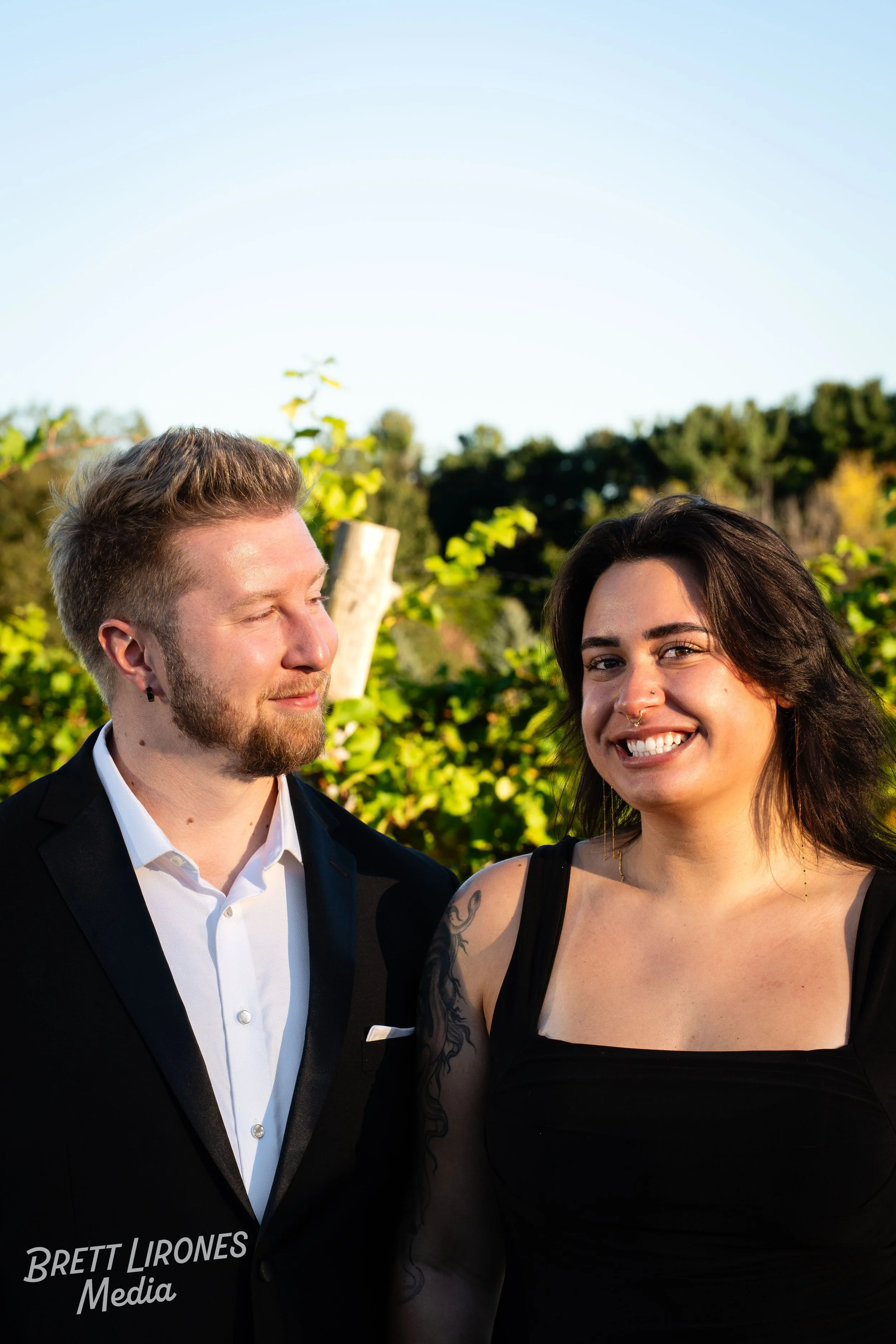A man in a black tuxedo and a woman in a black dress standing outdoors in a green, leafy setting with trees and a clear blue sky, smiling and looking at each other.