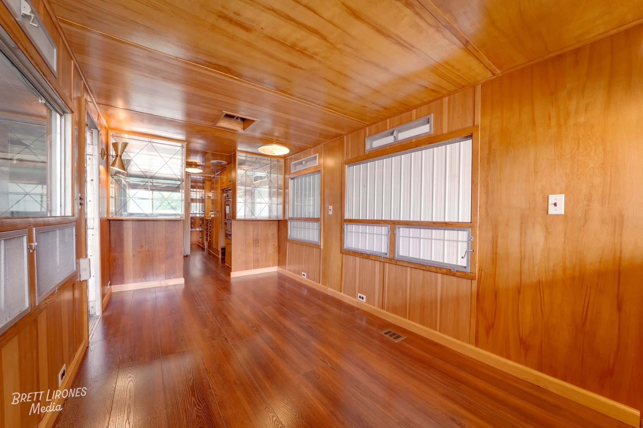 A room with wooden panel walls, ceiling, and floor, featuring multiple windows with white frames, some covered with blinds, and a visible ceiling vent and light fixtures.
