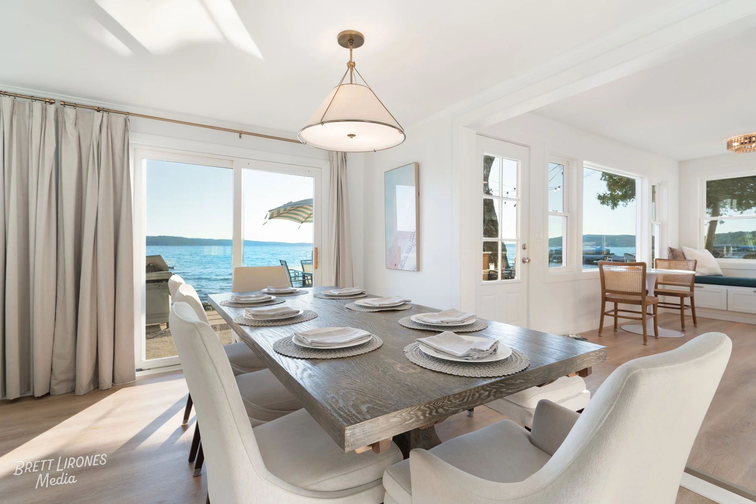 Dining room with a wooden table set with plates and napkins, overlooking a lake with an outdoor patio and beach umbrella visible through sliding glass doors.