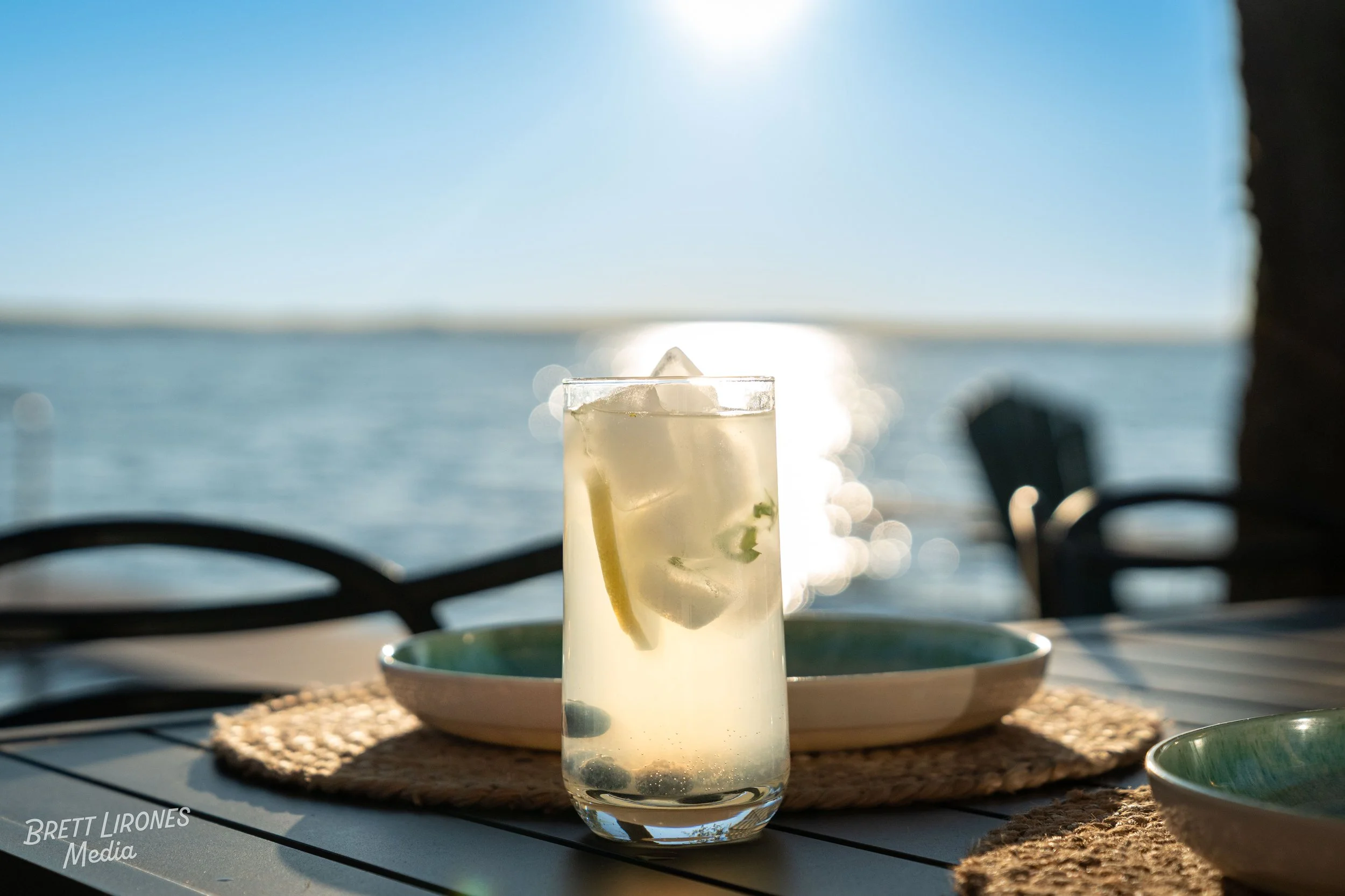 A glass of lemonade with lemon slices and ice cubes on a table outdoors near a body of water during sunset.