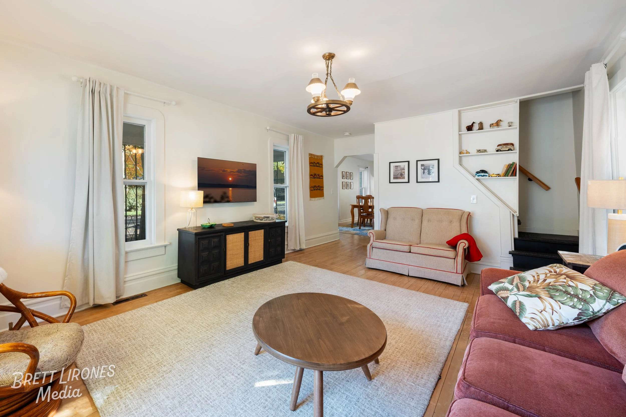 Living room with a beige sofa, a red blanket, a wooden coffee table, a TV mounted on the wall, windows with white curtains, and a hallway leading to other rooms.