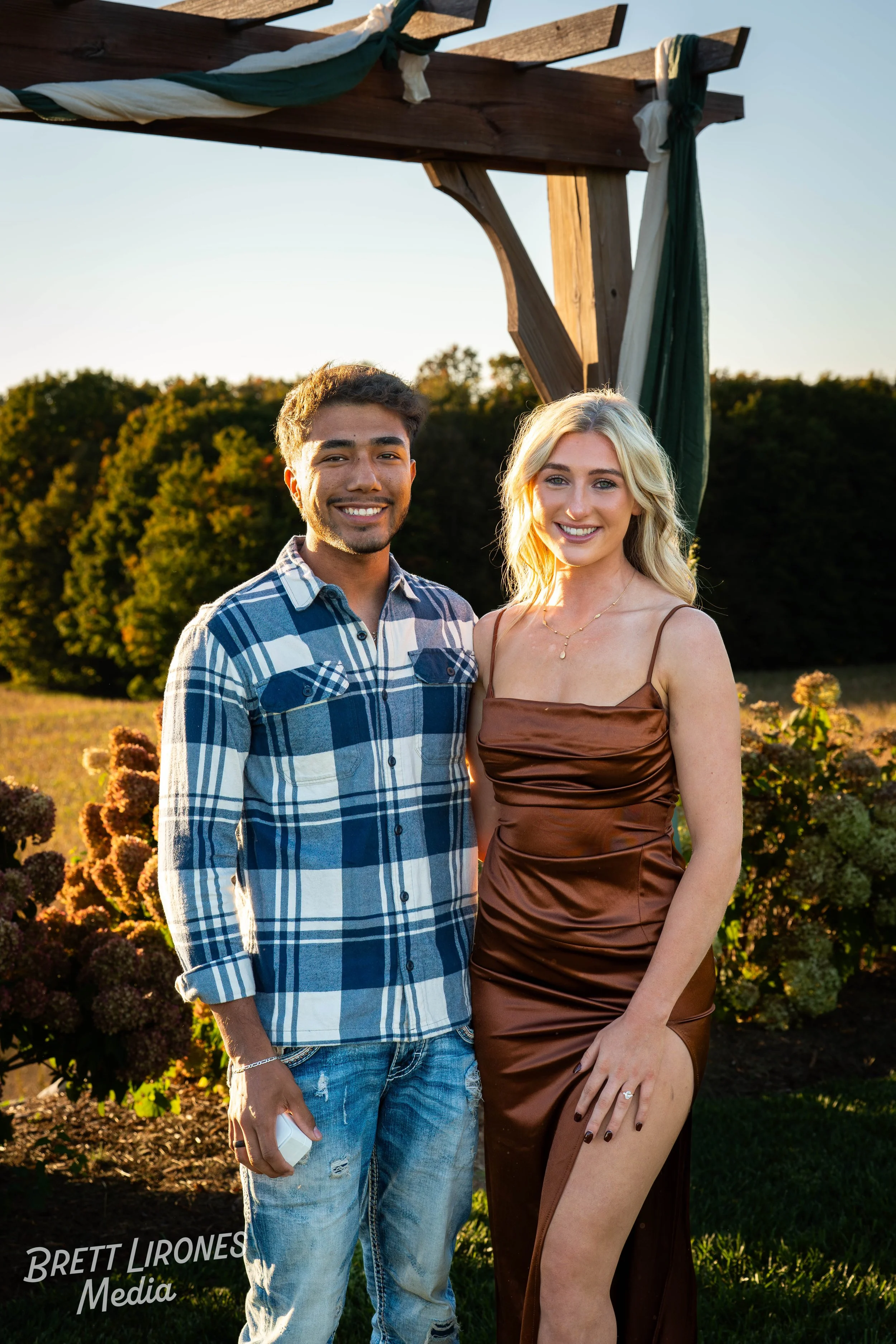 A young man and woman standing close together outdoors during sunset, smiling, in front of a wooden arch with greenery and bushes.