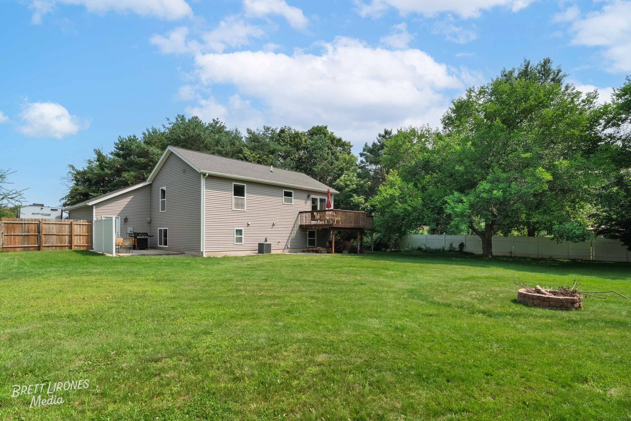 Backyard of a house with a grassy lawn, a large tree, a wooden deck with an umbrella, and a brick firepit, under a blue sky with white clouds.