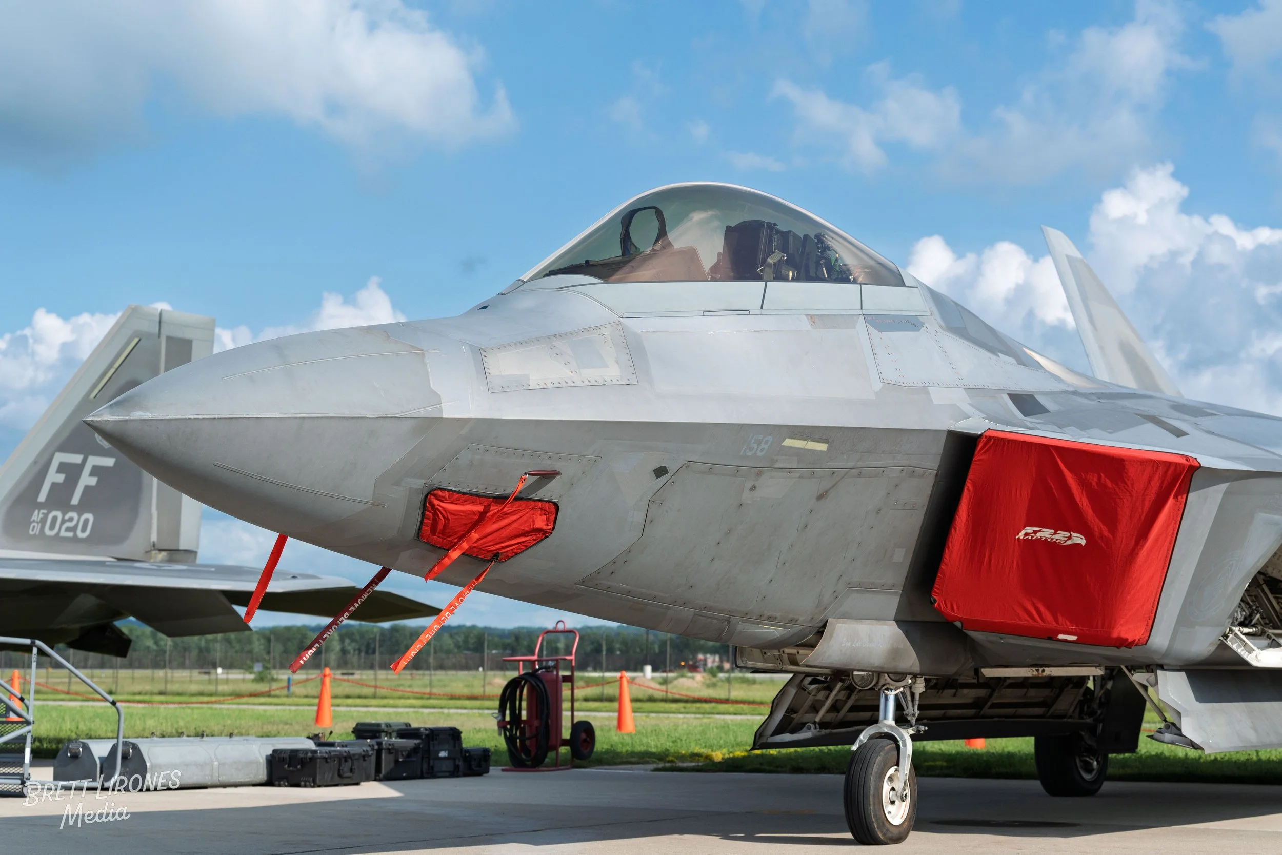 Close-up of a silver fighter jet aircraft parked on a tarmac with red covers over some openings, grass field and blue sky with clouds in the background.