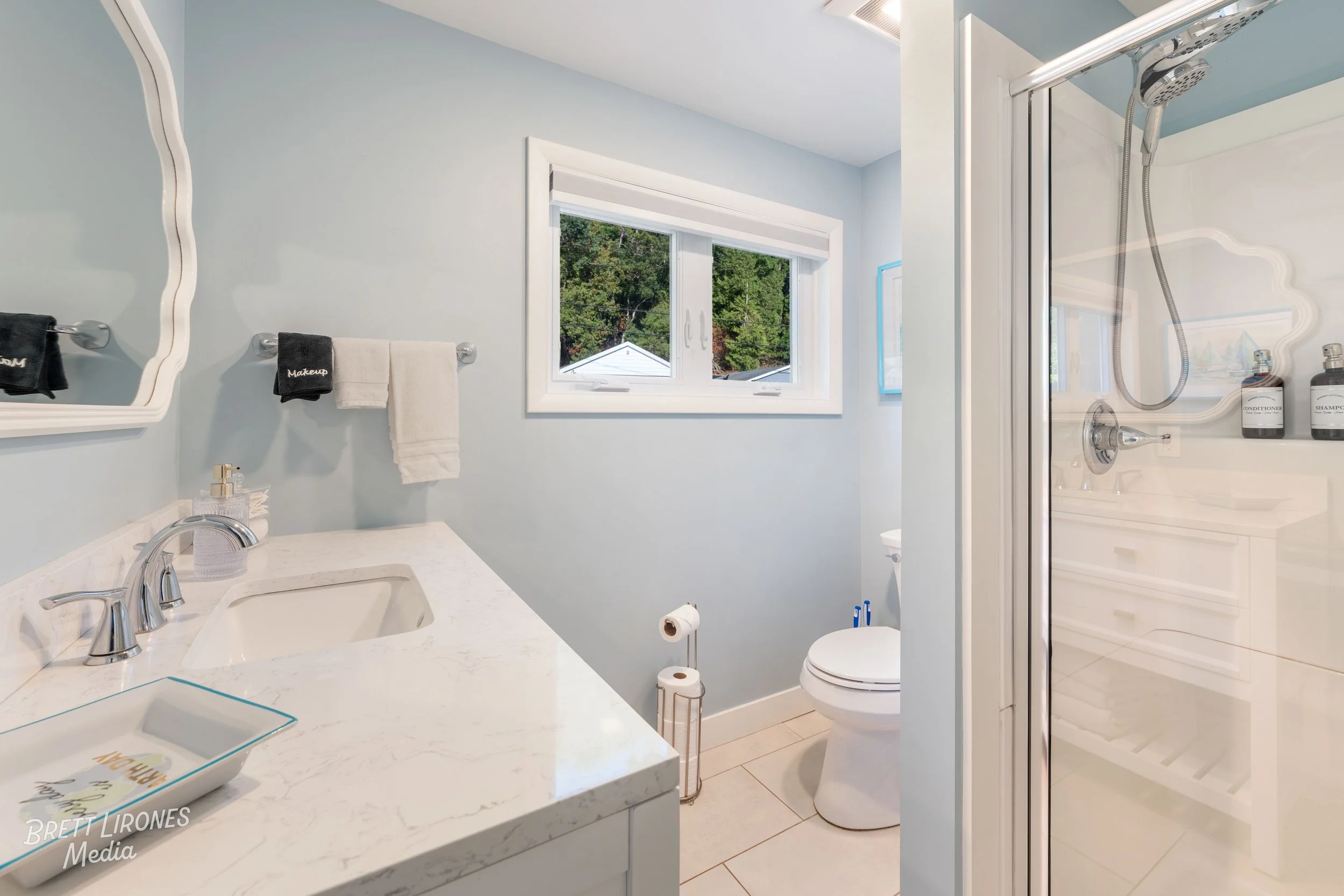 A clean, bright bathroom with light blue walls, a white countertop sink, a toilet, and a glass shower with labeled bottles inside. A window shows trees outside.
