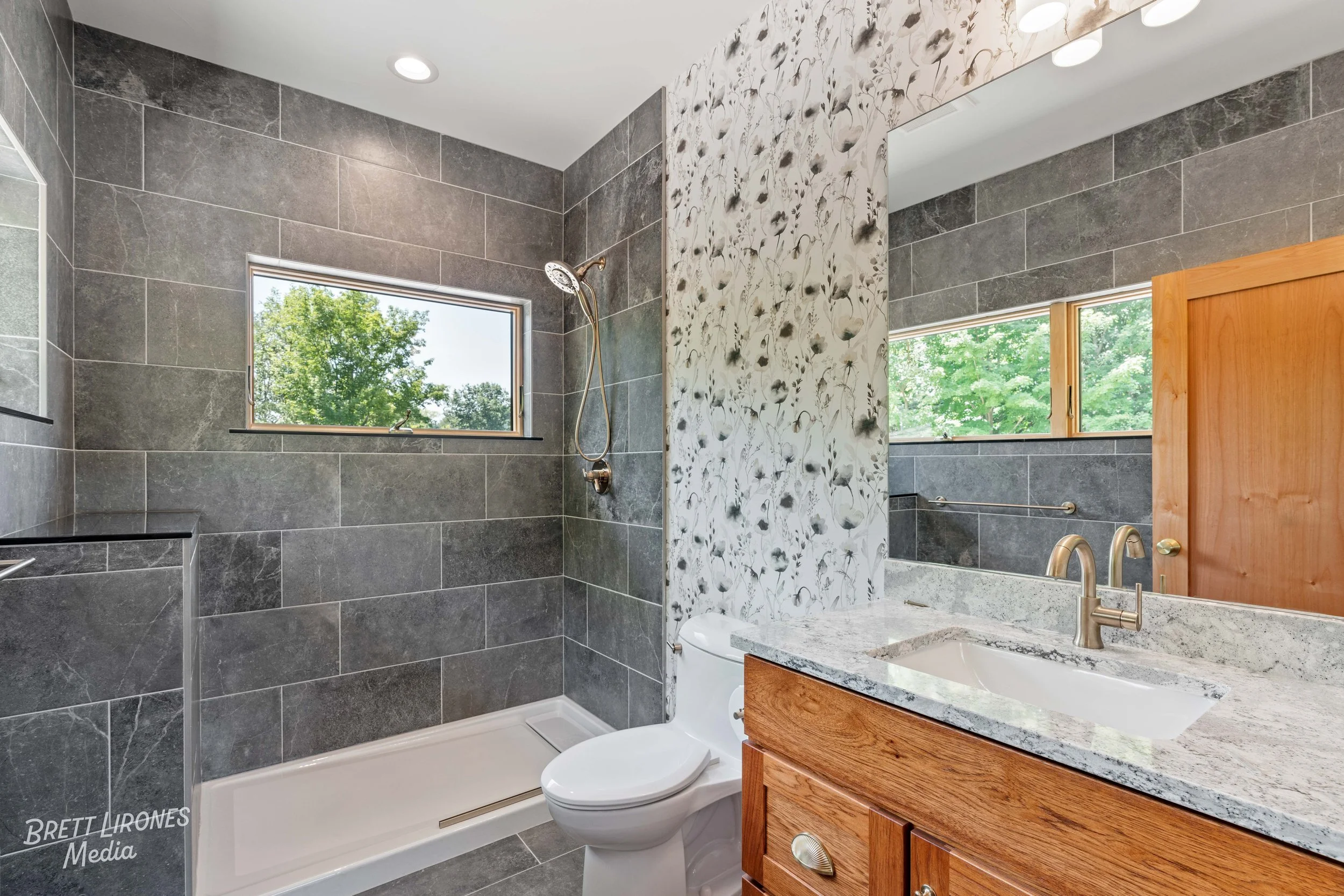 Bathroom with gray tiled walk-in shower, toilet, wooden vanity with granite countertop, large mirror, and window showing trees outside.
