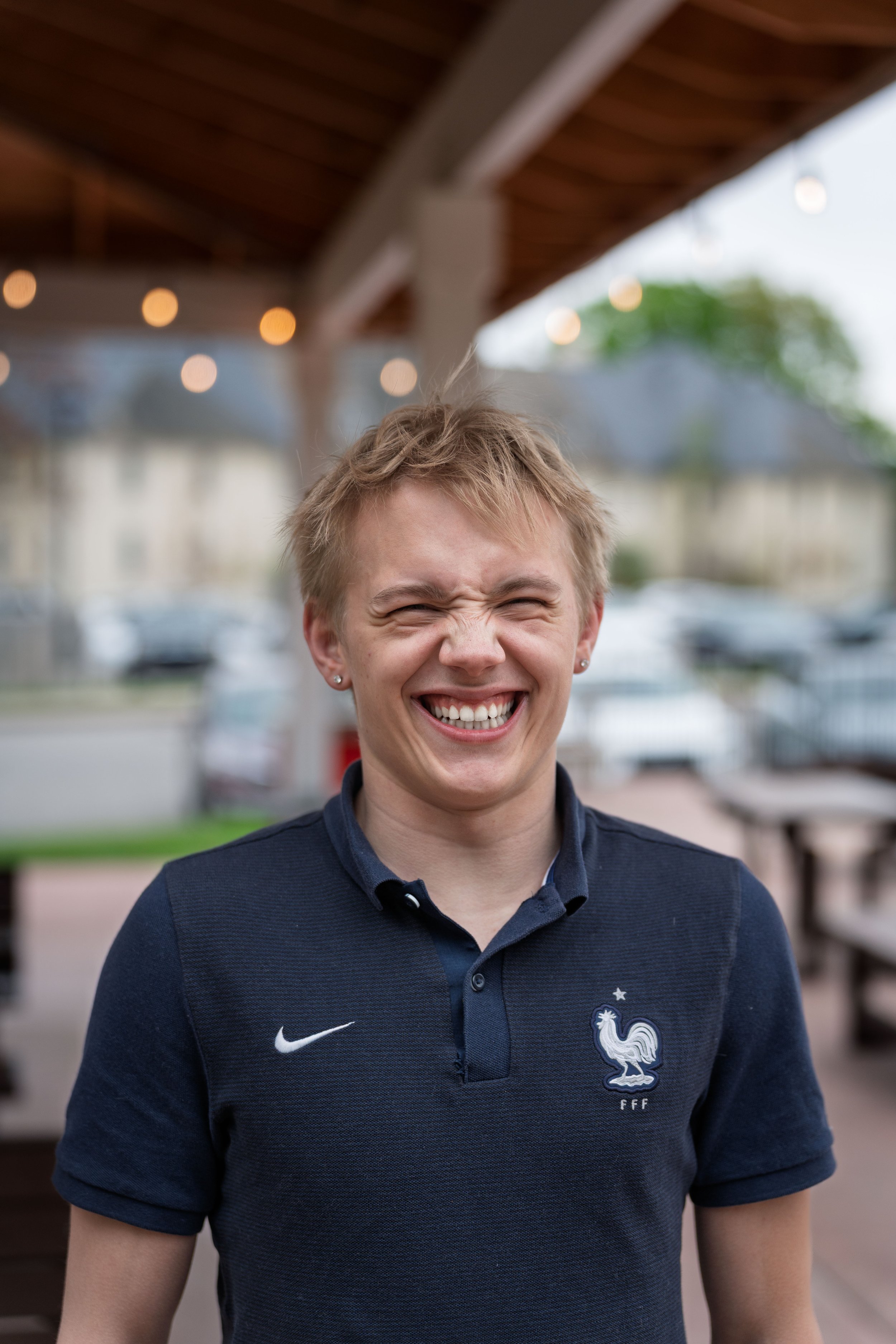 A young woman smiling widely, wearing a navy blue shirt with a French football team logo, standing outdoors with blurred background and string lights overhead.