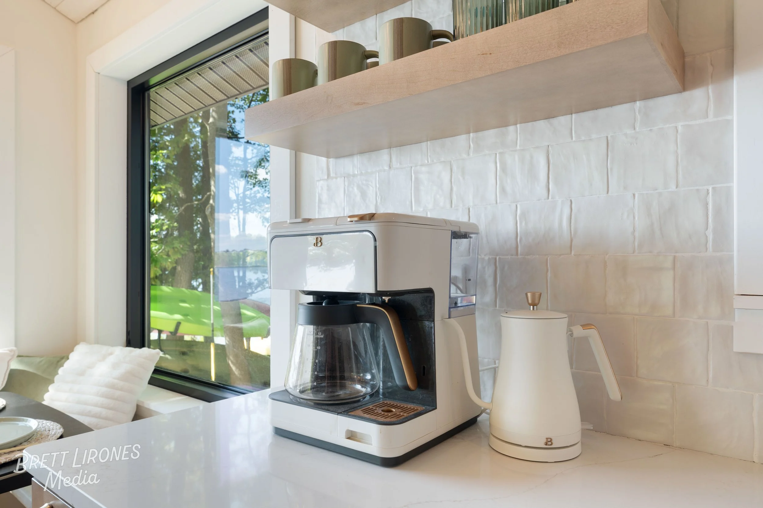 A white coffee maker and ivory electric kettle on a kitchen countertop near a large window with a view of greenery outside, with beige tiles on the wall behind.