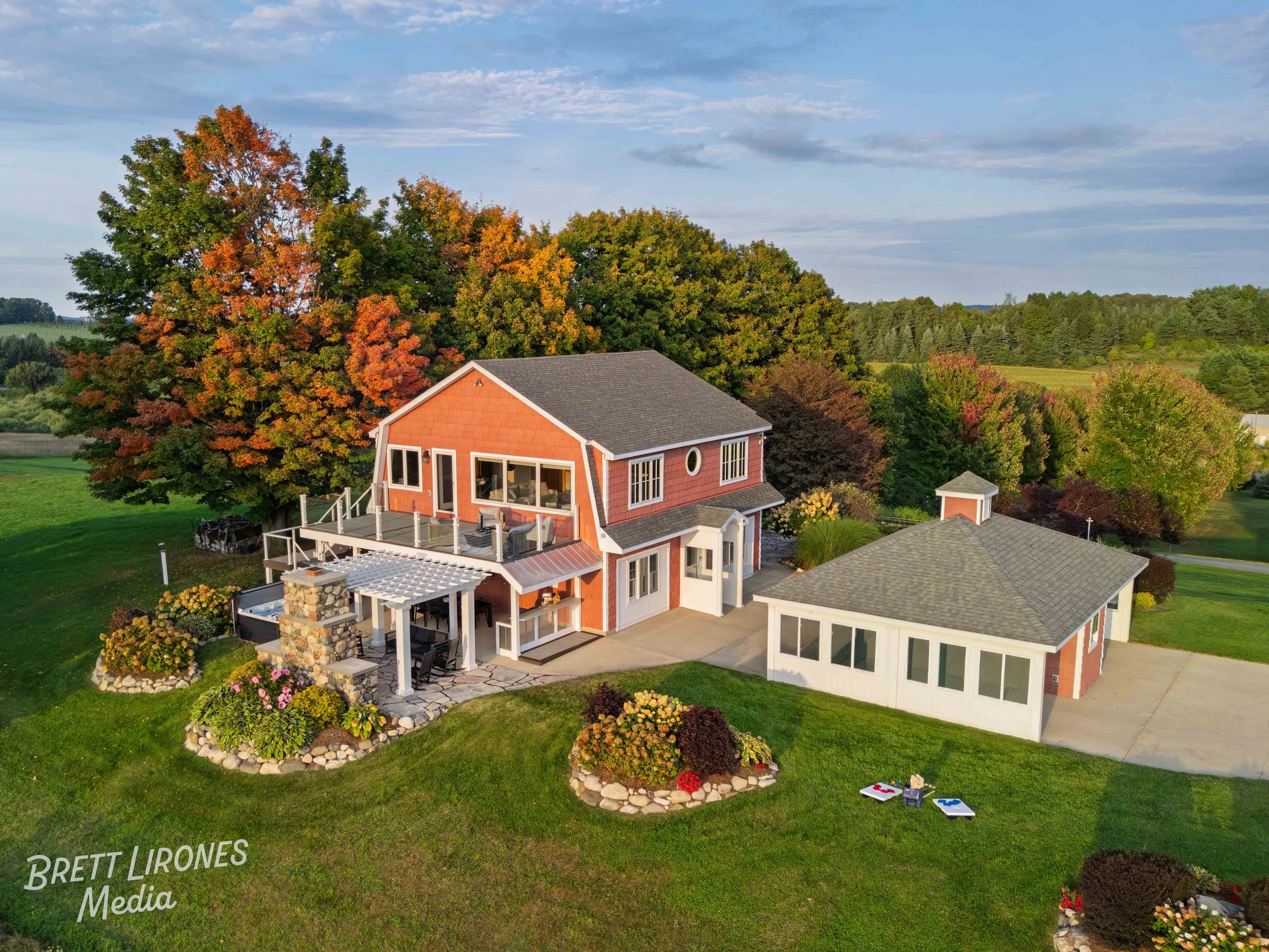 A house with a large deck and a detached garage is surrounded by green grass and trees with fall foliage. The property has landscaped flower beds, and there are cornhole boards on the lawn.