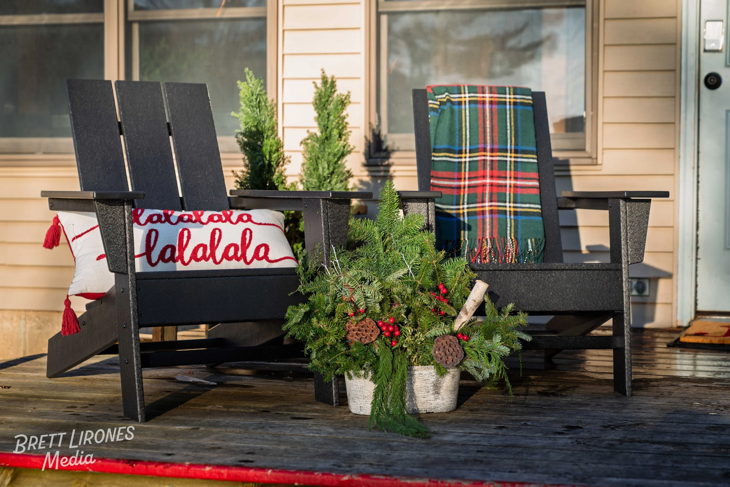 Black wooden porch bench with holiday decorations, including a red and white pillow with the text 'lalalala,' a plaid blanket draped over one side, and a white pot filled with fir branches, red berries, pinecones, and decorative items, on a wooden de