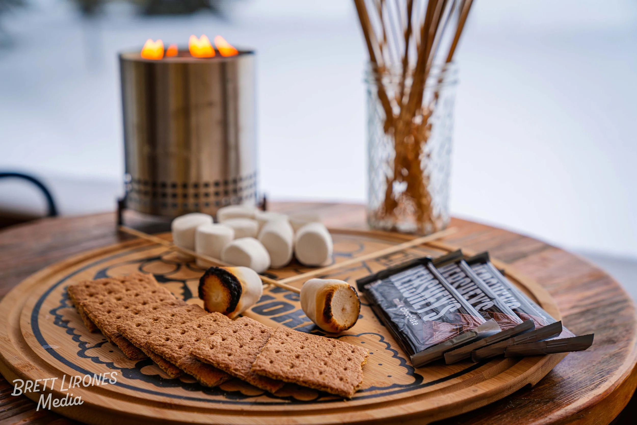 A wooden tray with graham crackers, marshmallows, and chocolate bars, with a metal burner and a glass vase with sticks in the background.