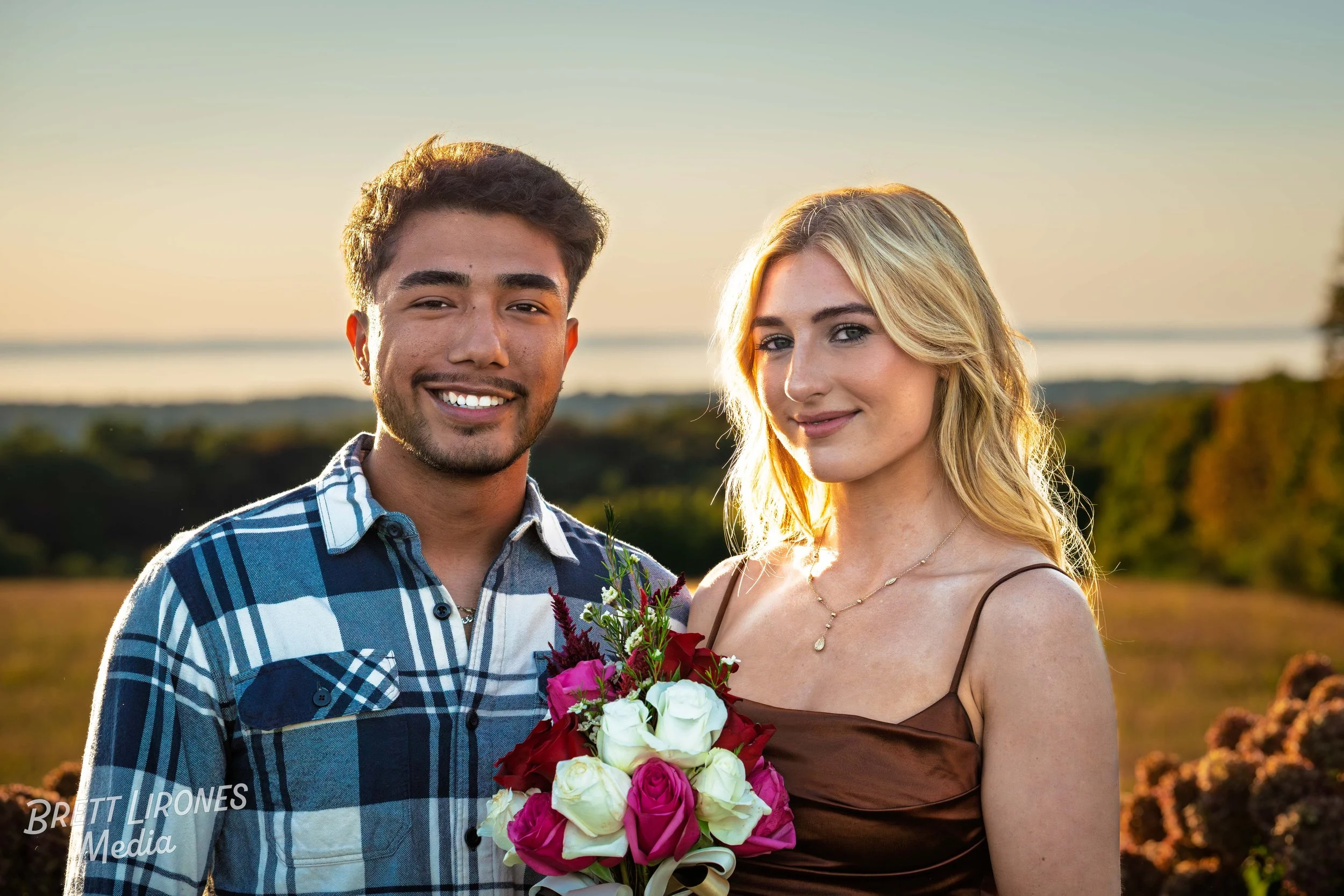 A couple standing outdoors during sunset, with the man smiling and wearing a plaid shirt and the woman holding a bouquet of pink, white, and red roses, wearing a brown dress.