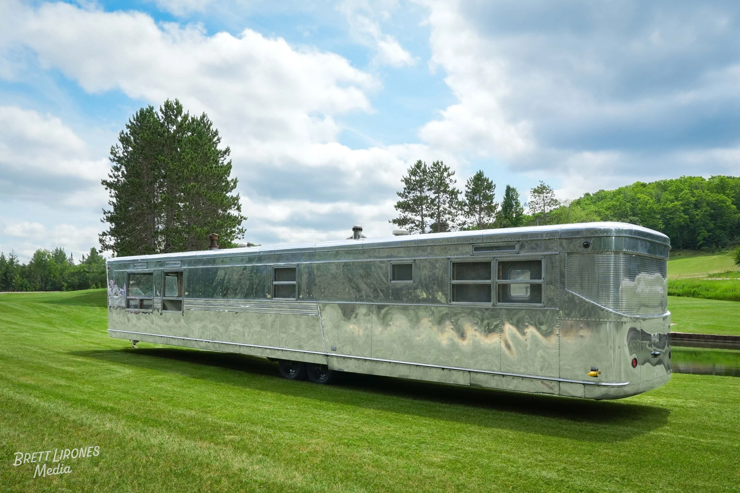 A shiny, metallic camper trailer parked on a lush green field with trees in the background and a partly cloudy sky overhead.