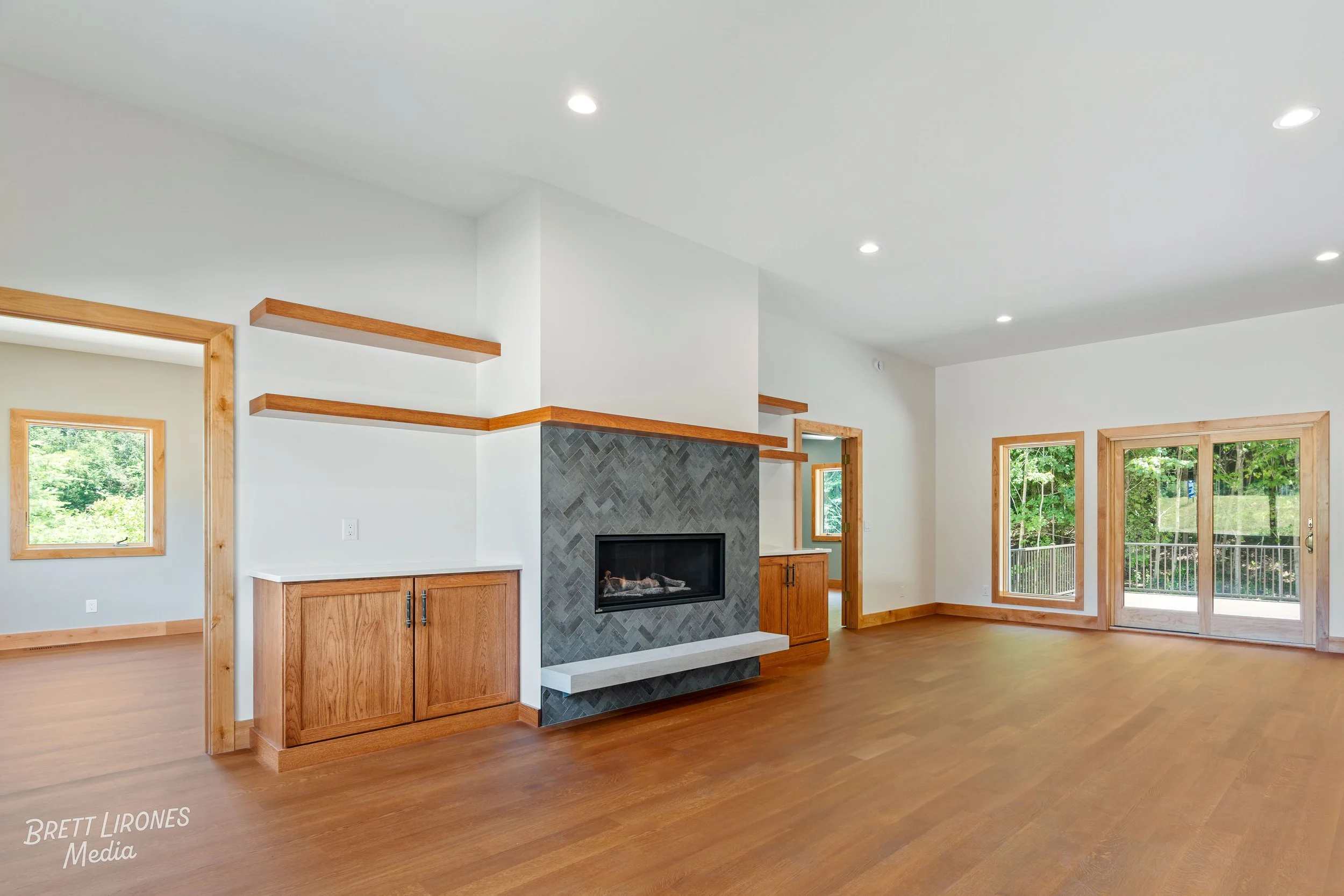 Empty living room with wood floors, a modern fireplace with gray tiles, and large windows and sliding glass door leading to outside, with wooden trim and built-in wooden cabinets.