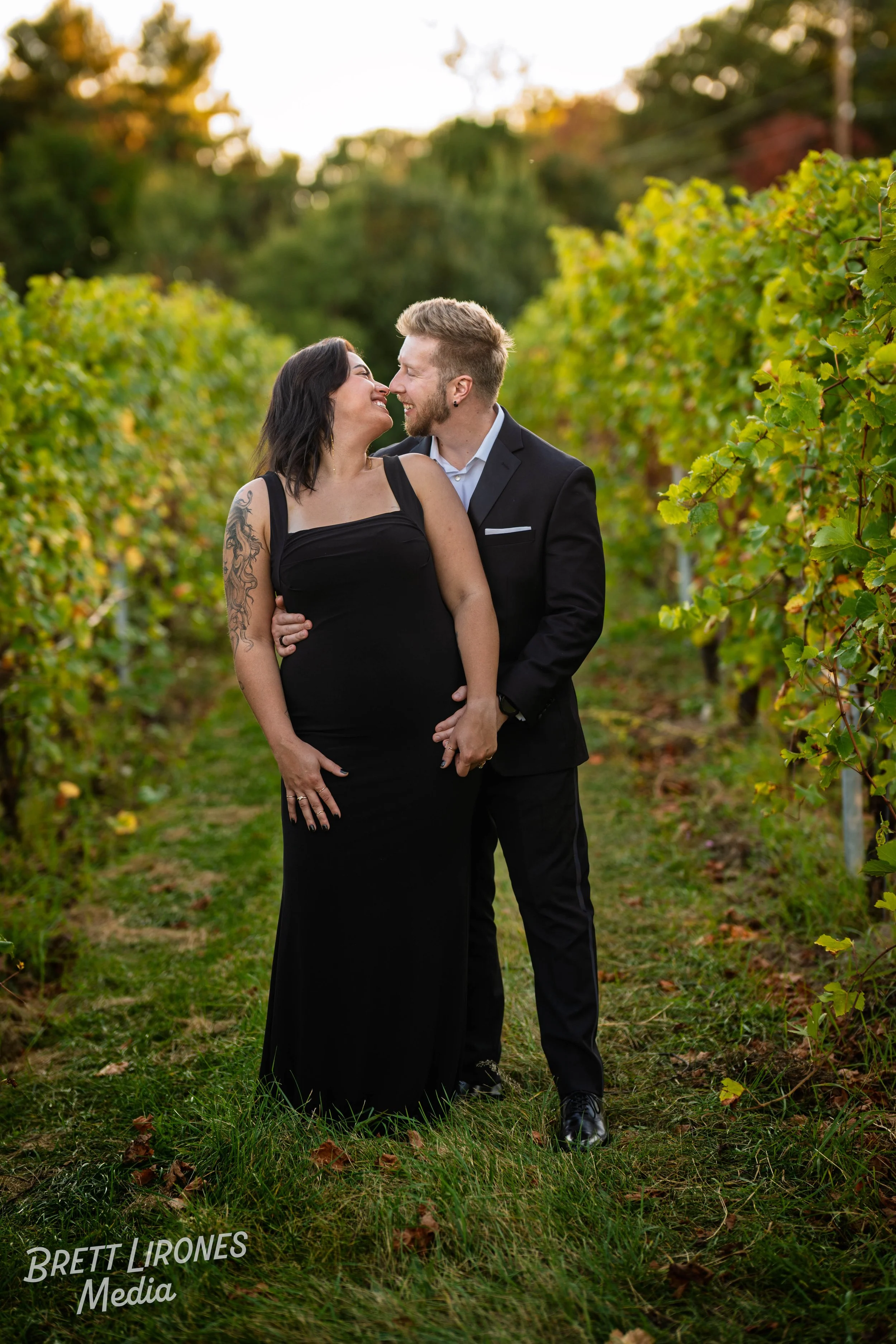 A couple in formal attire kissing in a vineyard during sunset, with lush grapevines on either side.