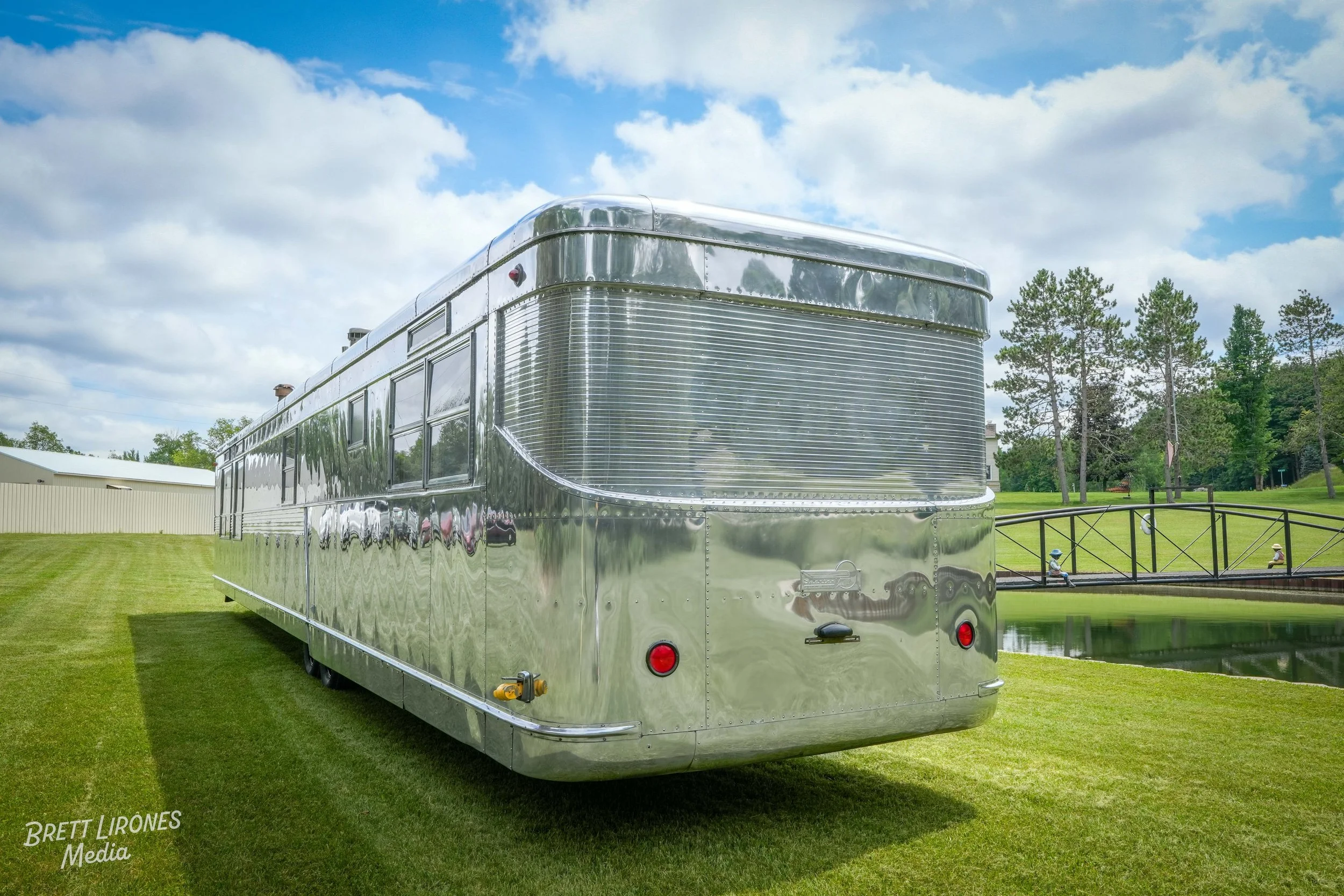 A shiny, silver vintage trailer parked on a grassy area near a pond with a small bridge, surrounded by trees, under a partly cloudy sky.