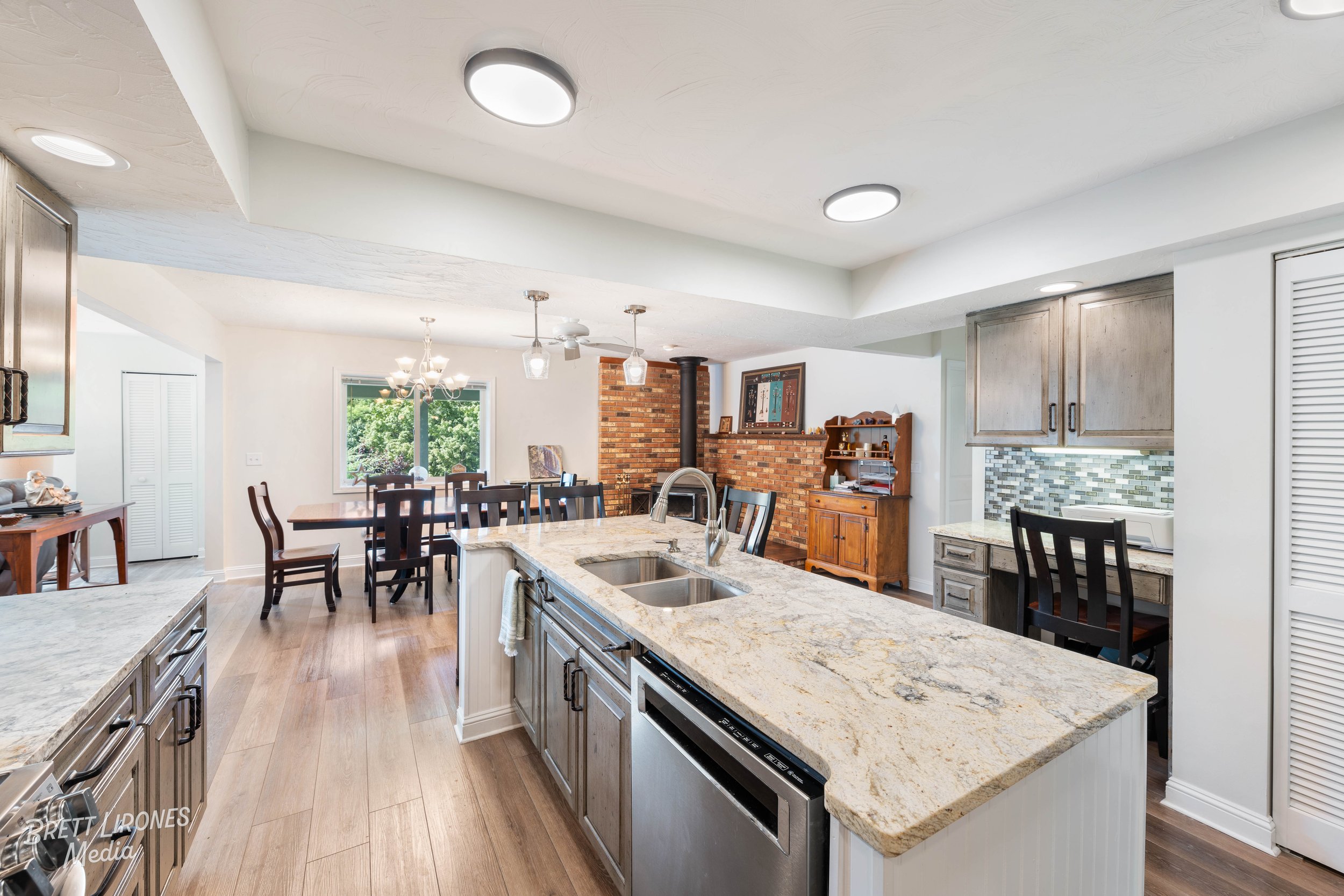 Open-concept kitchen and dining area with granite countertops, wooden cabinetry, stainless steel appliances, a brick fireplace, and a dining table with black chairs, illuminated by natural light from a large window.
