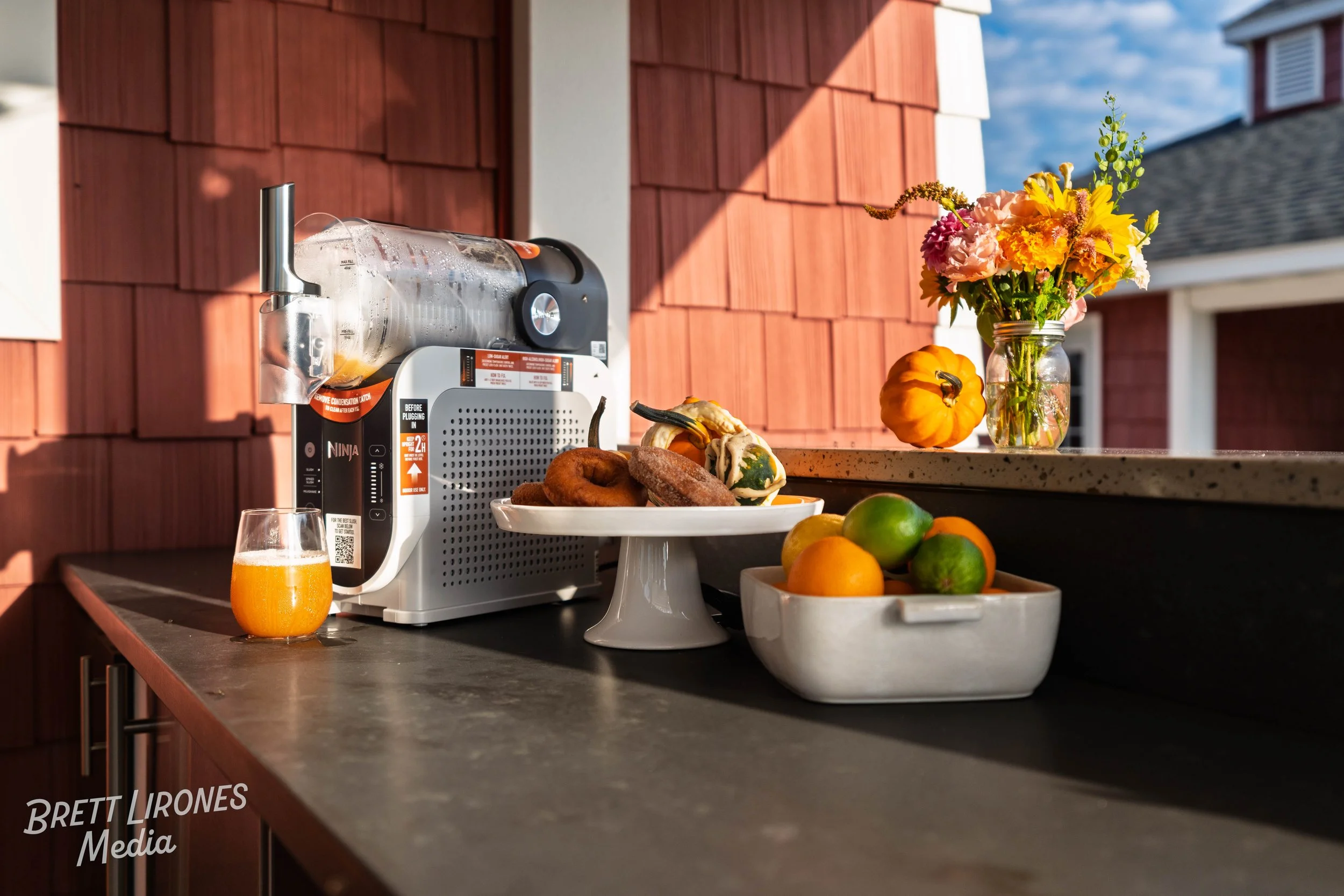 Outdoor kitchen counter with a juice machine, glass of orange juice, a white cake stand with donuts and gourds, a white bowl with oranges and limes, and a glass jar with pink and yellow flowers and small pumpkin.