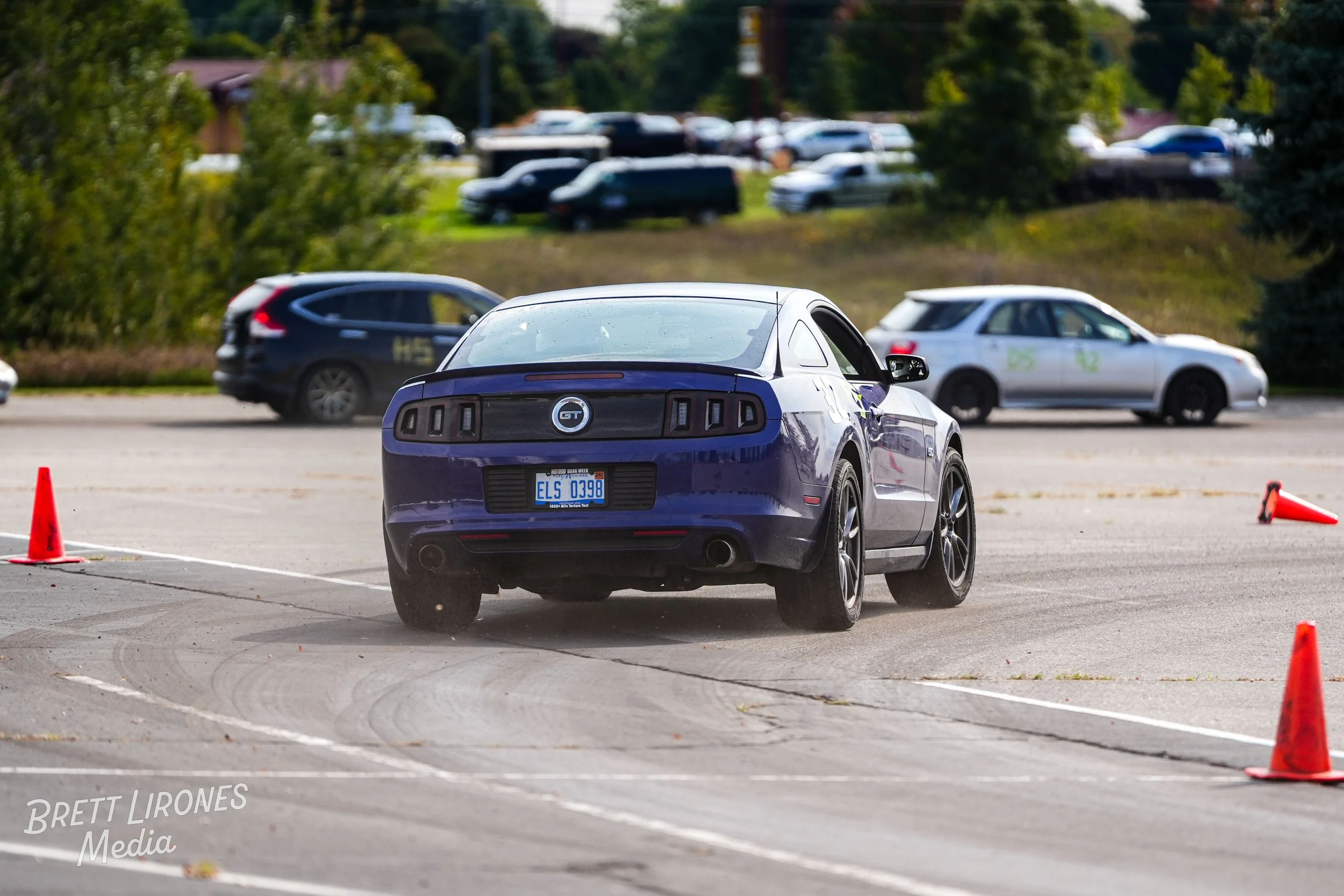 A purple Ford Mustang GT driving through an autocross course with orange cones in a parking lot, with other parked cars and greenery in the background.