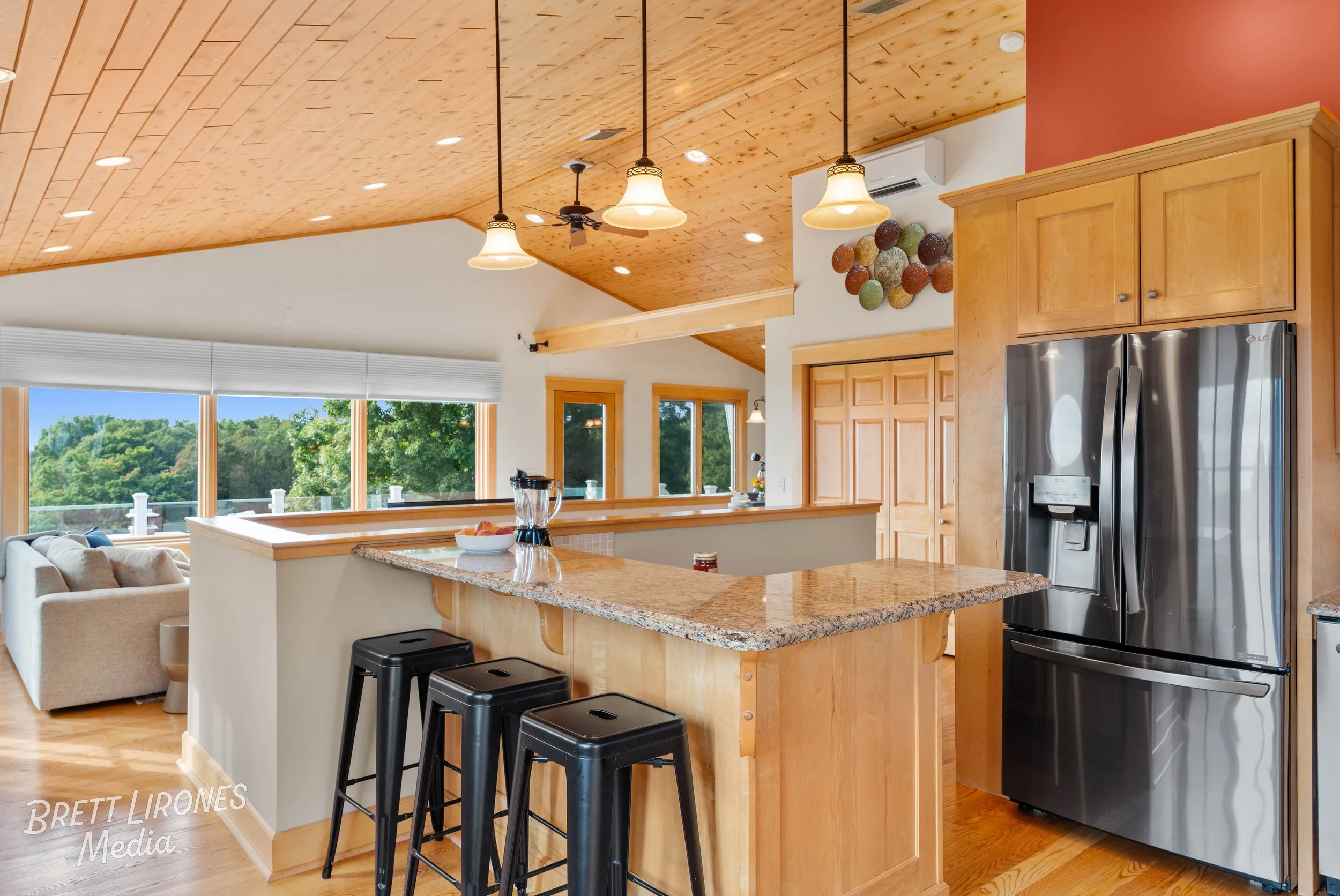 Kitchen with wooden cabinets, stainless steel refrigerator, granite countertops, and three black barstools. Open living space with large windows showing green trees outside, wooden ceiling with pendant lights, and a beige couch in the background.
