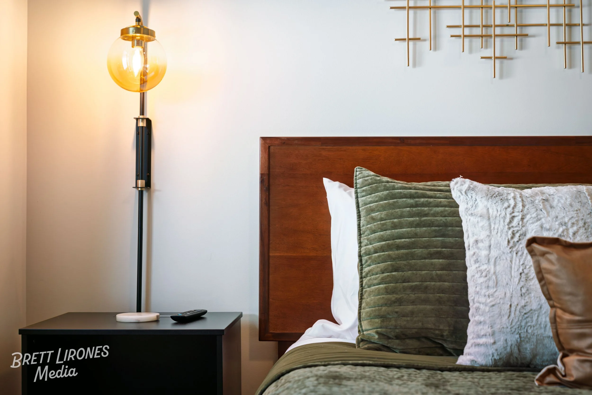 Close-up of a bed with green and white textured pillows, a wooden headboard, and a side table with a lamp, remote, and small device in a bedroom.