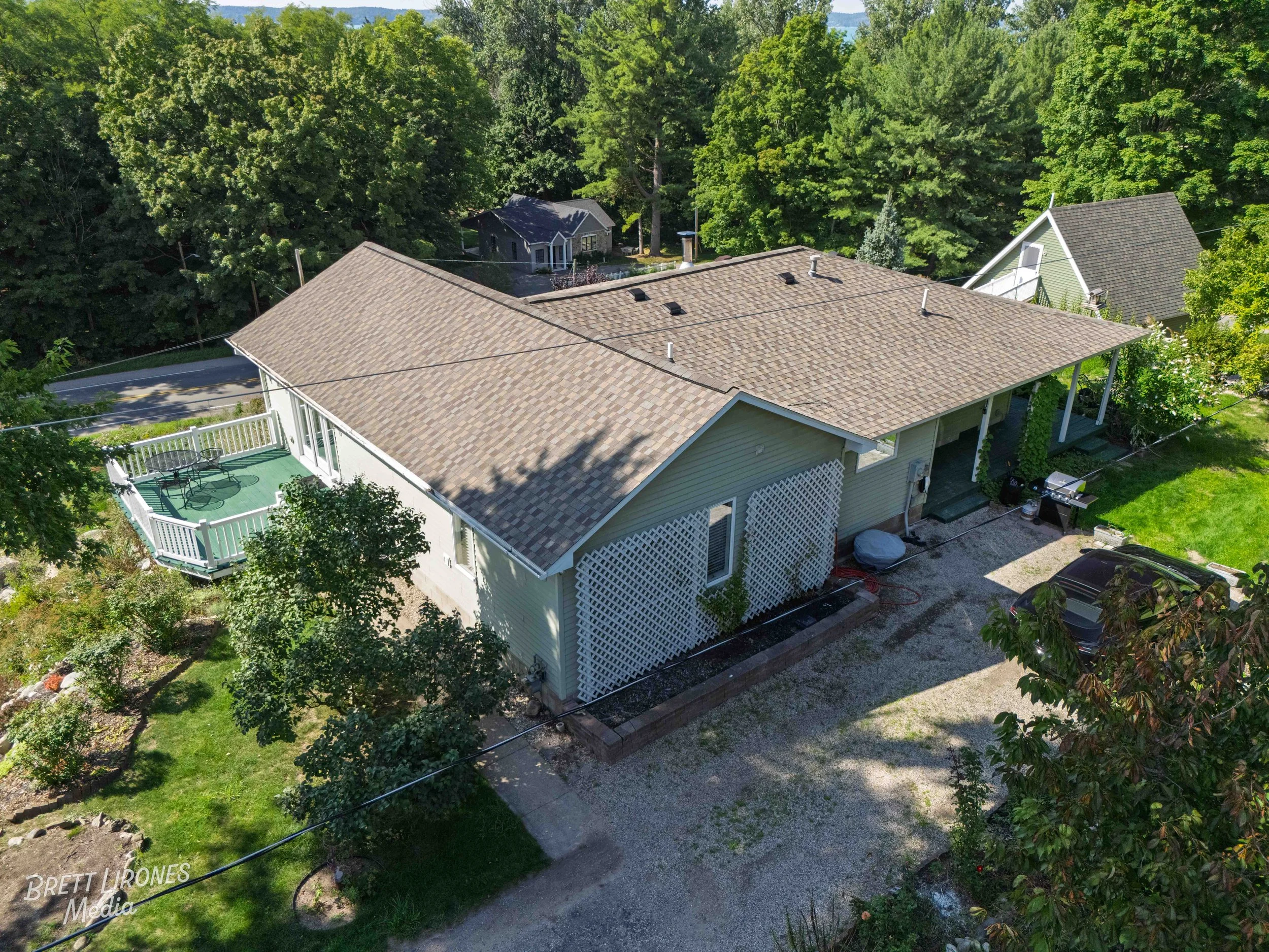 Aerial view of a house with beige roof, white siding, a small deck with furniture, and a gravel driveway with a black car parked. Surrounded by trees and neighboring houses.