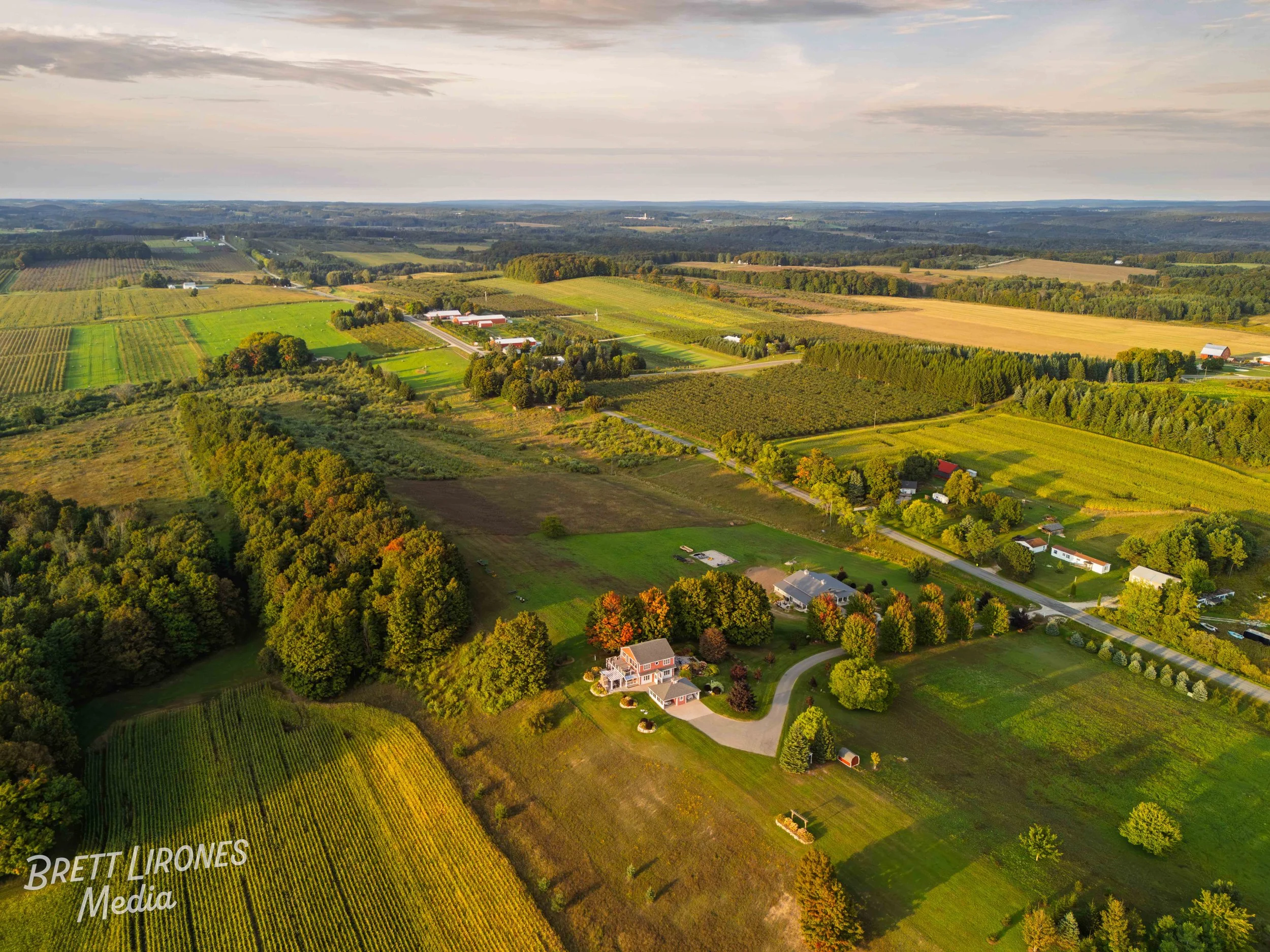 Aerial view of a rural landscape with houses, trees, farmland, and fields under a cloudy sky at sunset.
