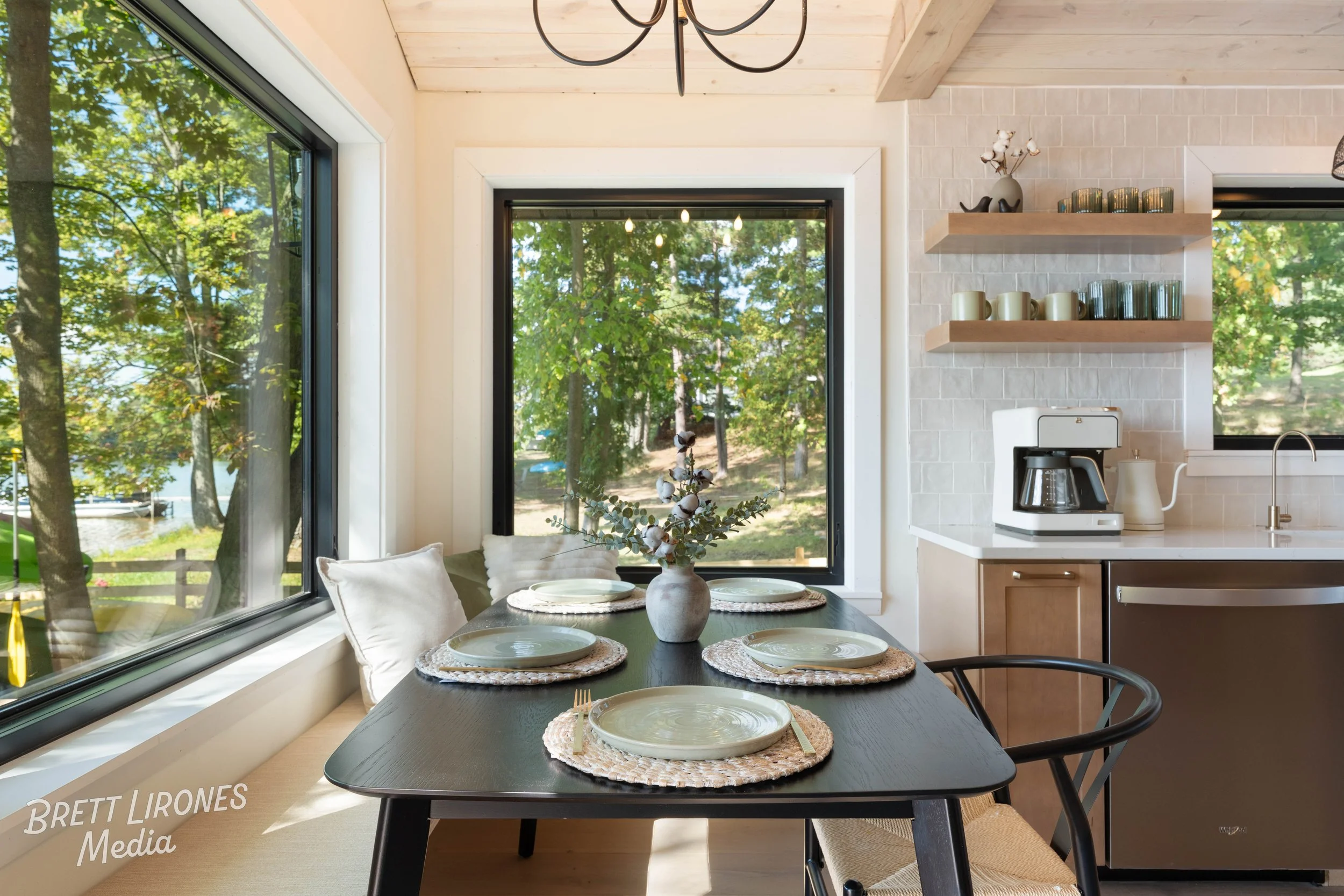 A dining area with a dark wooden table set with four woven placemats, ceramic plates, and cutlery, positioned next to large windows showing a view of green trees outside. The room features light-colored walls, a white ceiling with exposed wood beams,