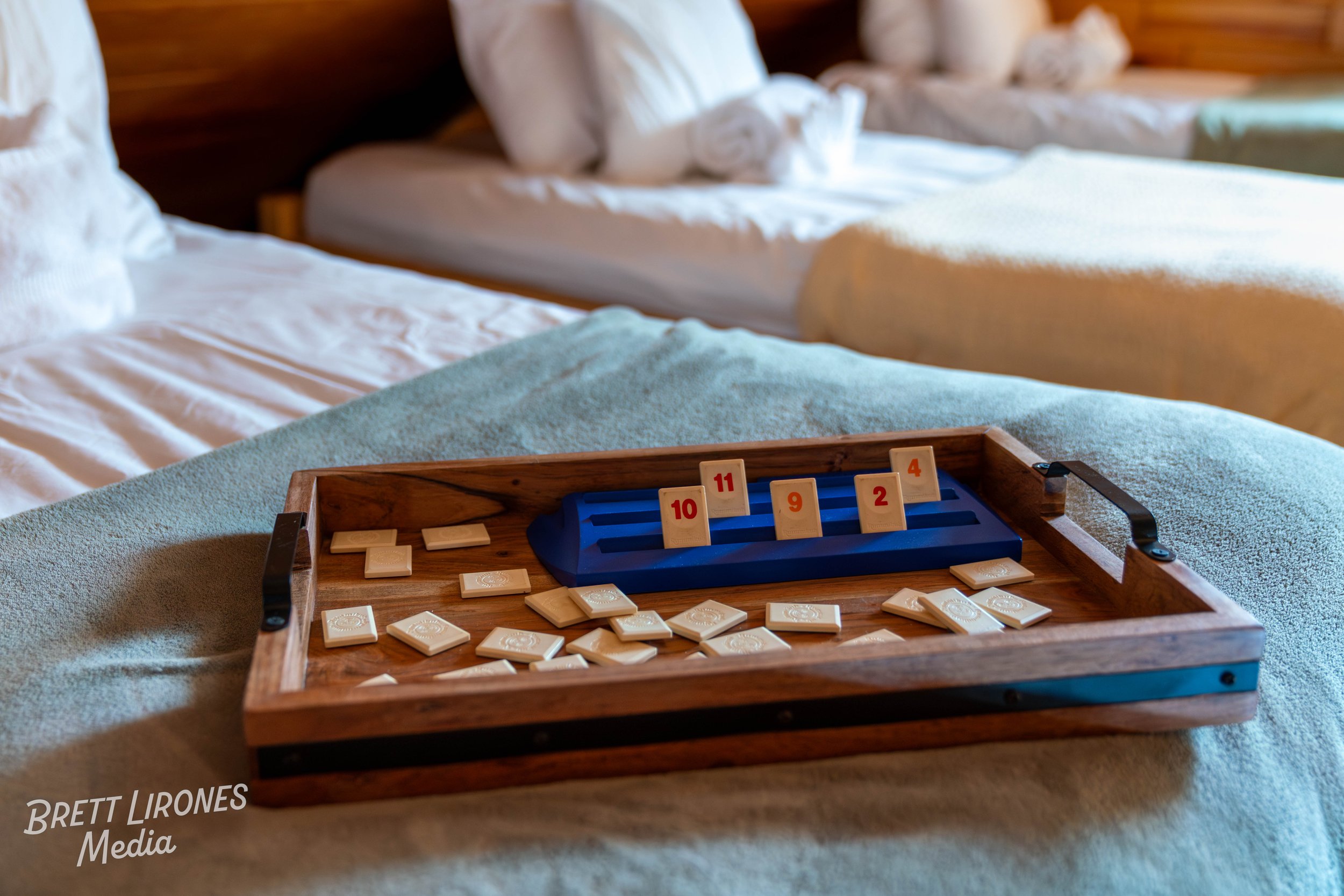 A wooden tray with a tropical-themed design, containing a game of Rummikub on a bed with white sheets and towels rolled into the pillows. The game tiles are scattered on the tray and the table.