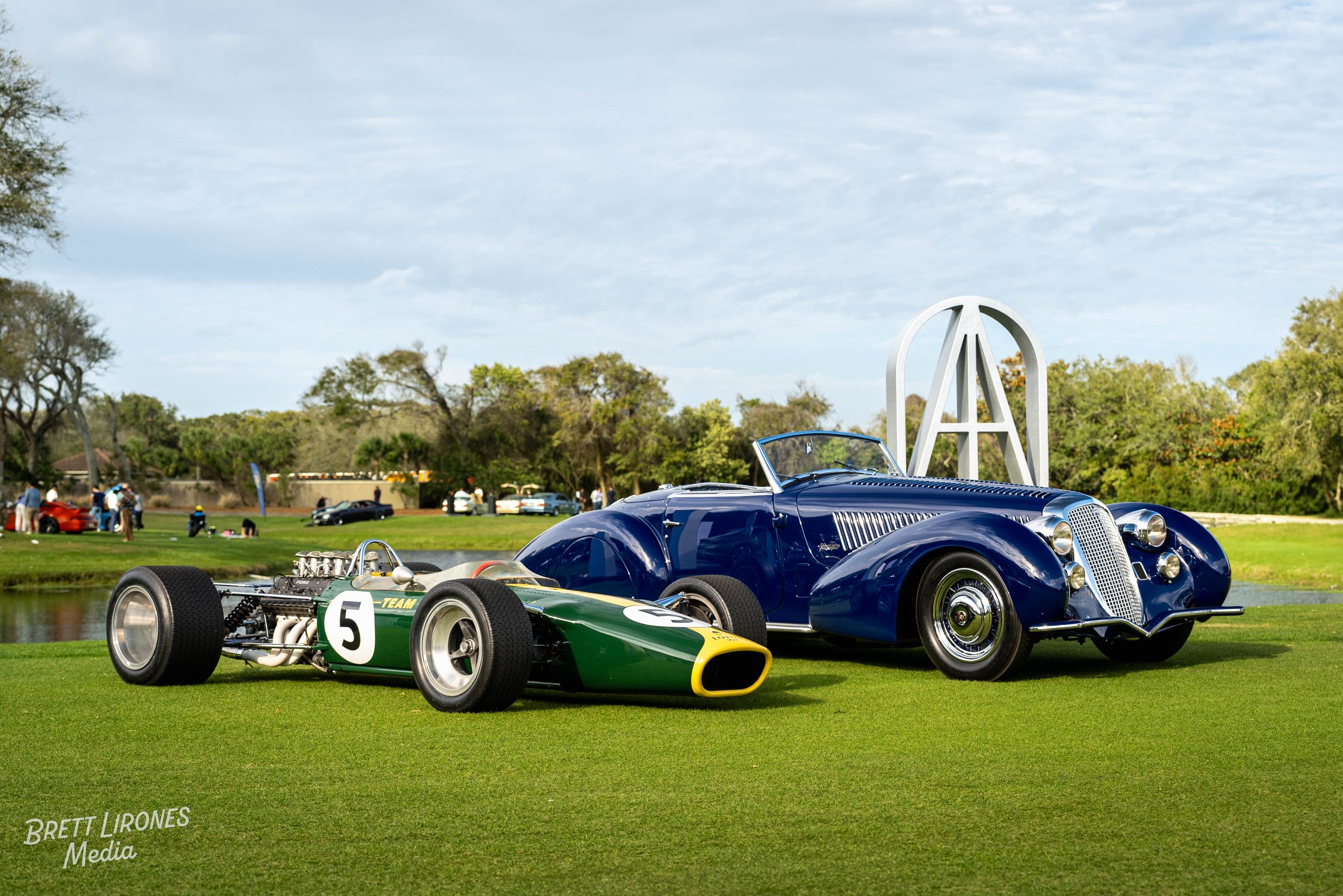 A vintage green and yellow race car with the number 5 on it and a classic blue car with a prominent grille and curved fenders parked on a grassy area near a water hazard at a car show, with trees and spectators in the background.