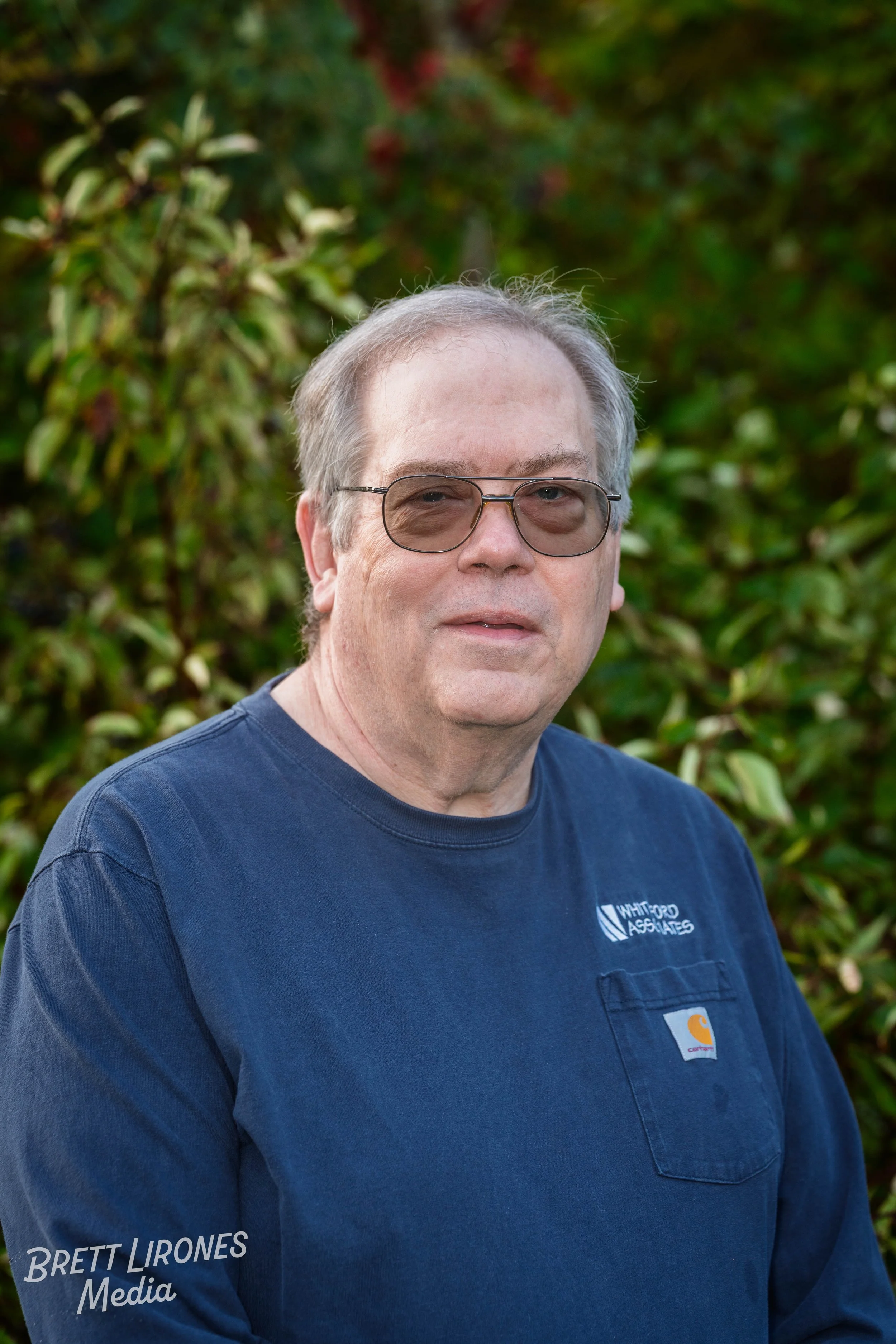 An older man with gray hair wearing glasses and a navy blue Carhartt shirt with a logo on the chest, standing outdoors with green foliage in the background.