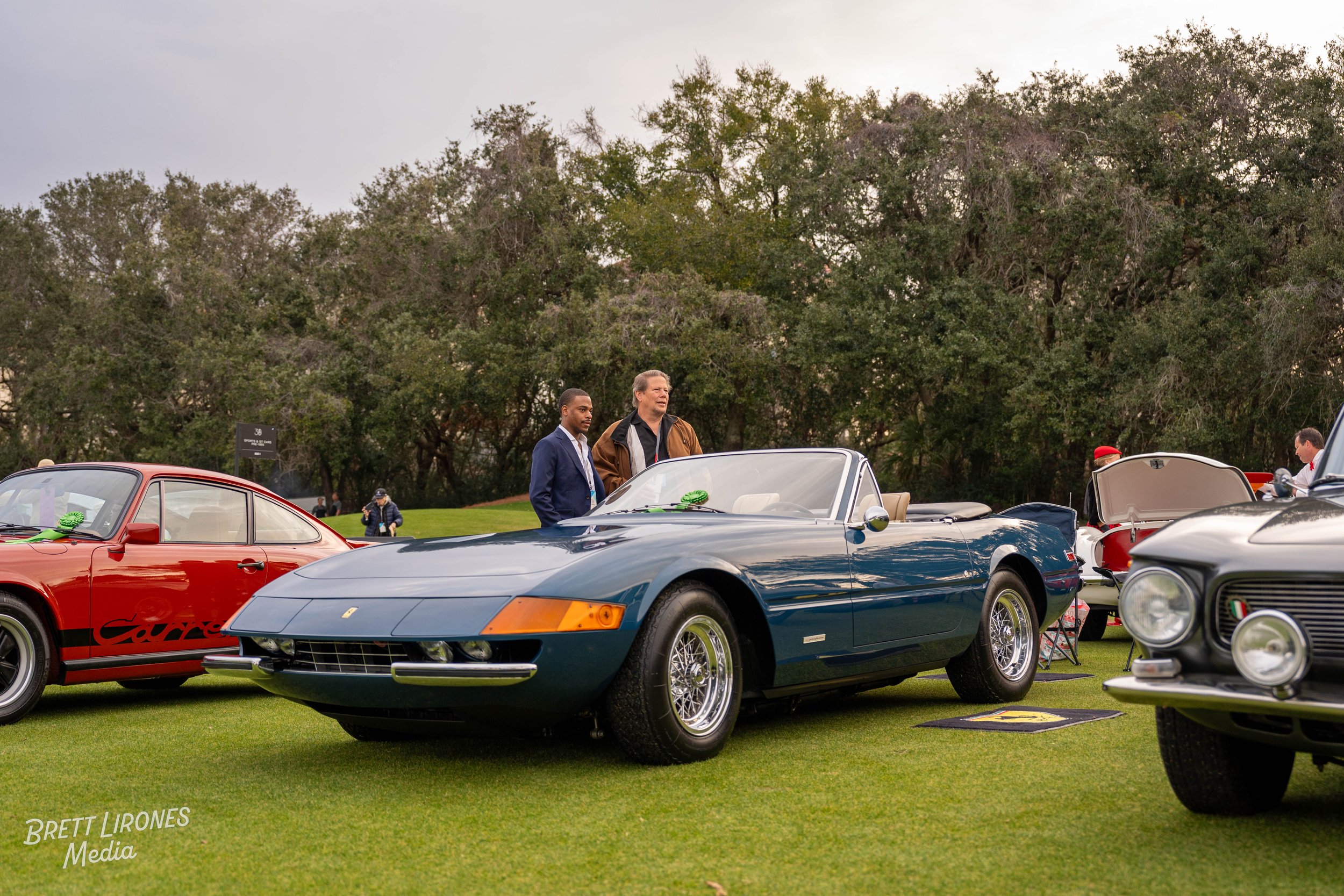A vintage blue Ferrari sports car parked on the grass at a car show, with two men looking at it. Other classic cars and trees are visible in the background.