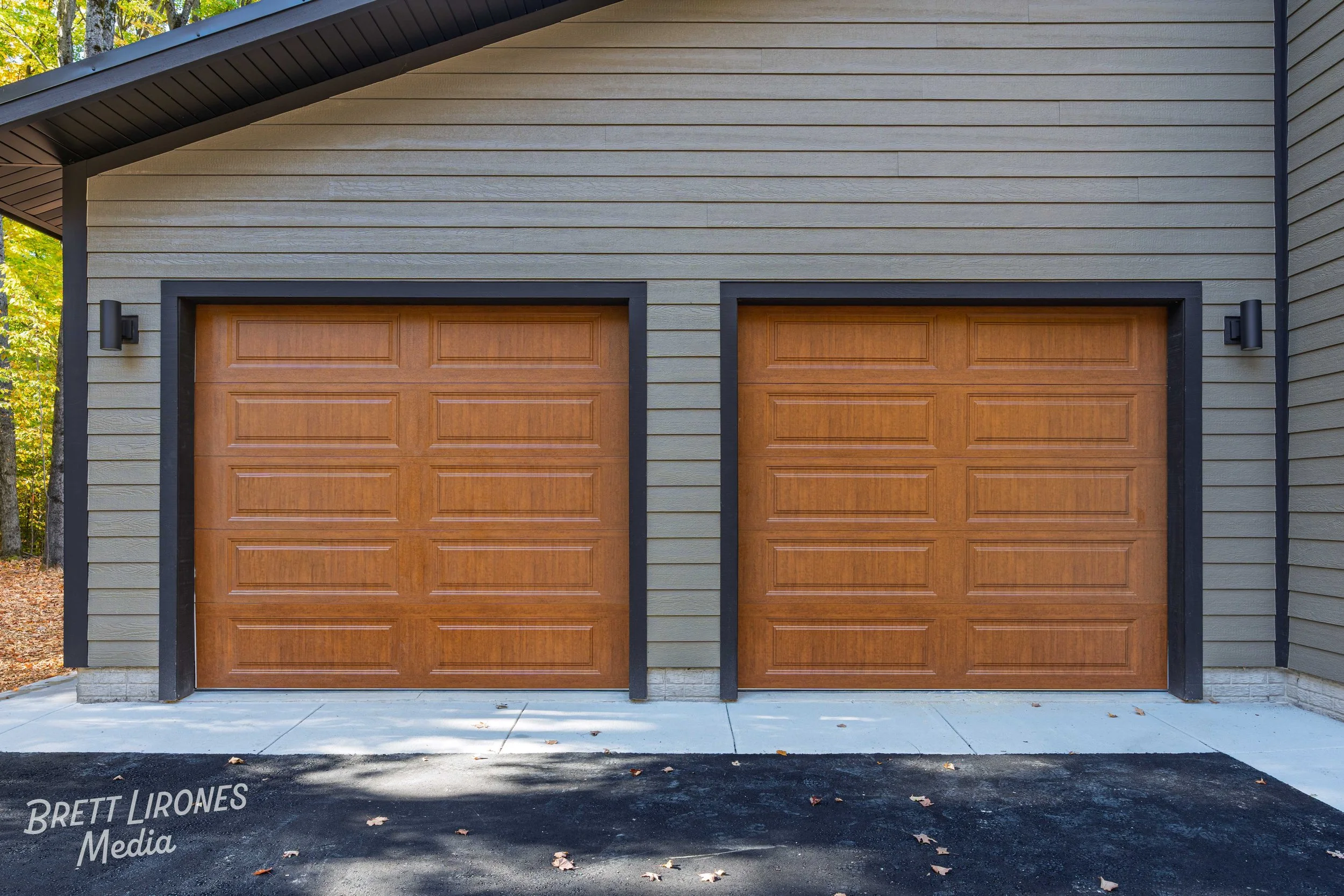 Two wooden garage doors with black trim on a house with beige horizontal siding, black light fixtures on each side, and a concrete driveway with scattered leaves.