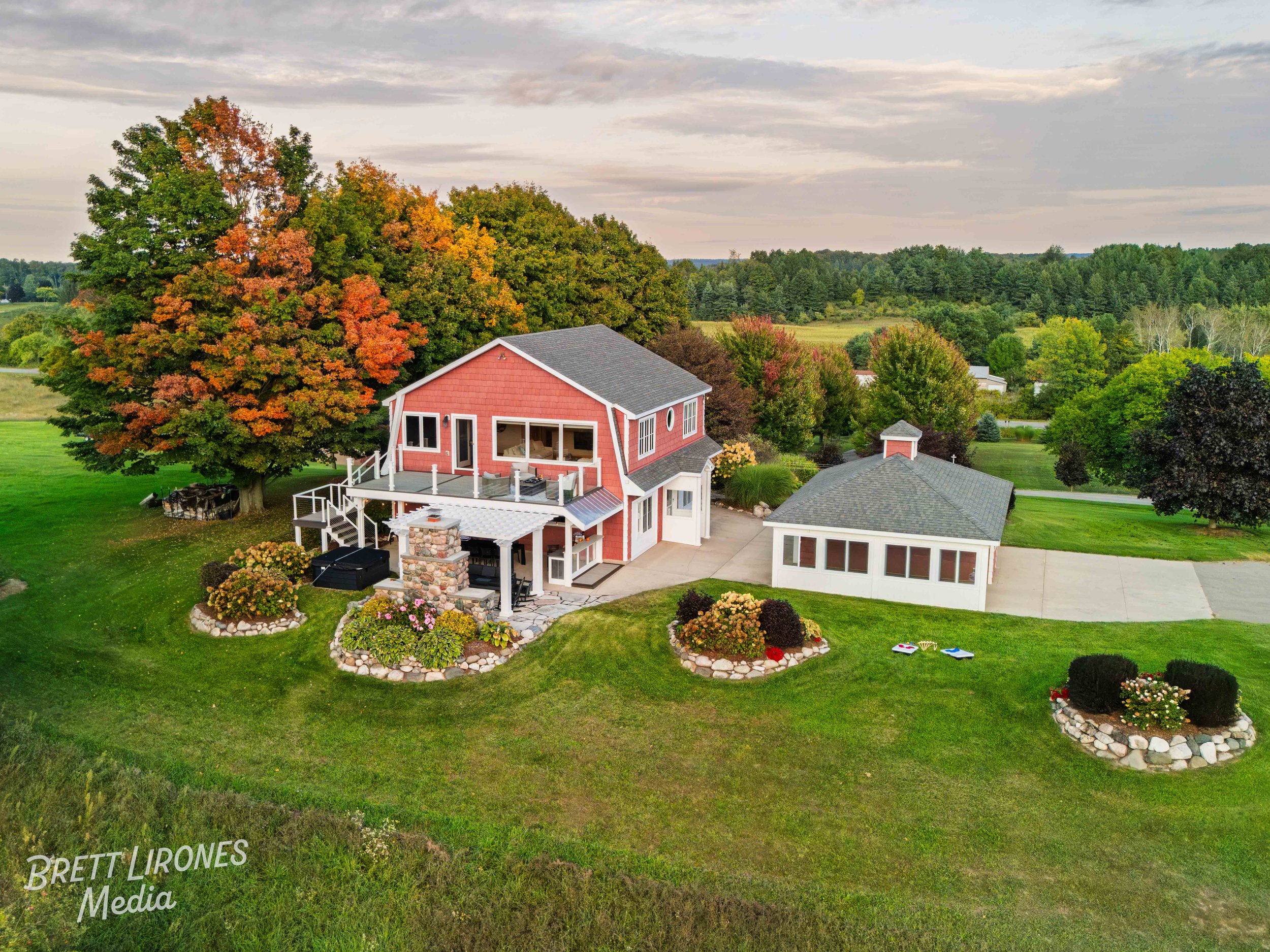 A large two-story red house with a porch, surrounded by colorful autumn trees and a well-maintained lawn with landscaped flower beds, a detached garage, and a stone patio with outdoor furniture, all set in a rural landscape.