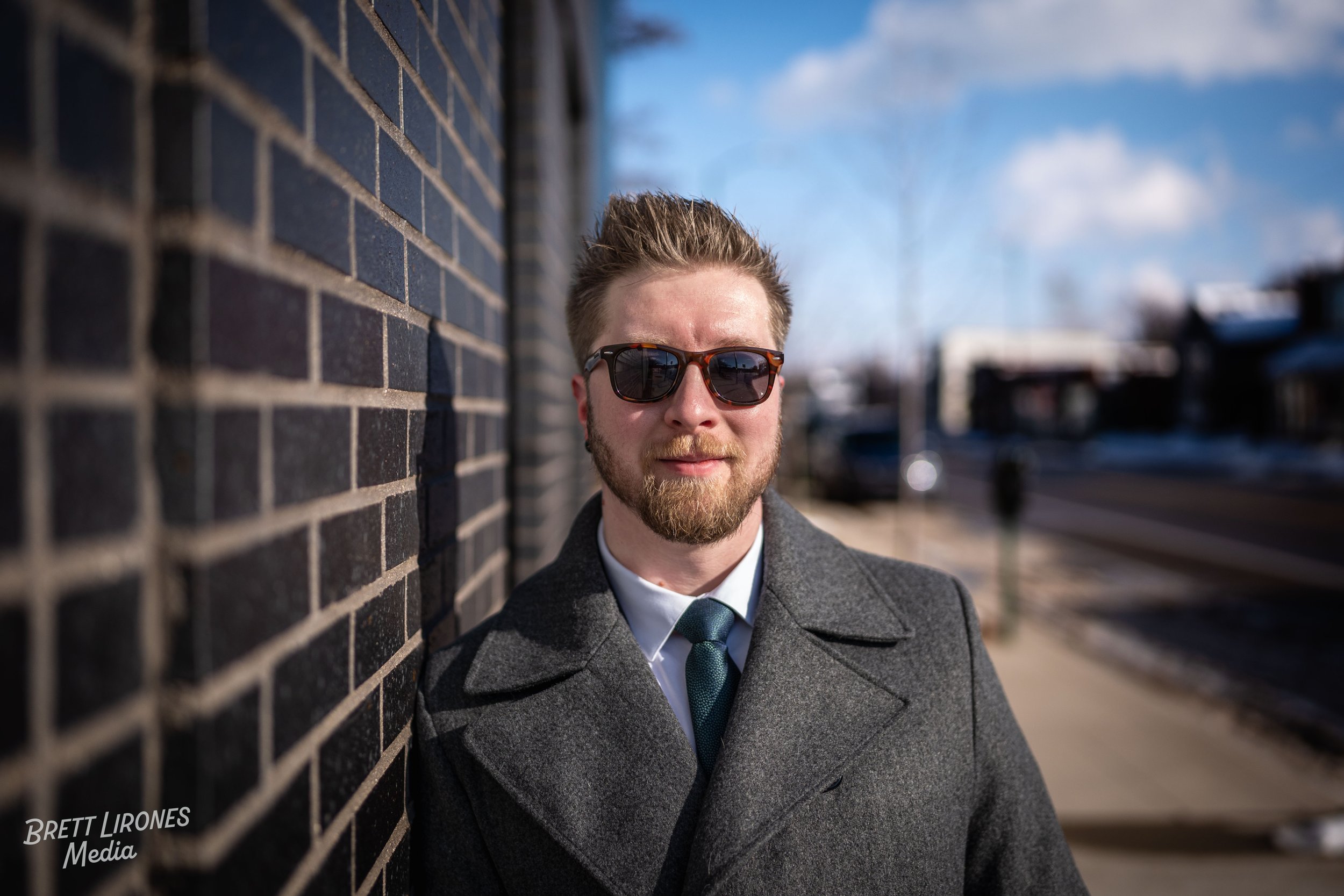 A man with sunglasses, a beard, and styled hair wearing a gray coat and tie, leaning against a brick wall on a sunny day in an urban area.