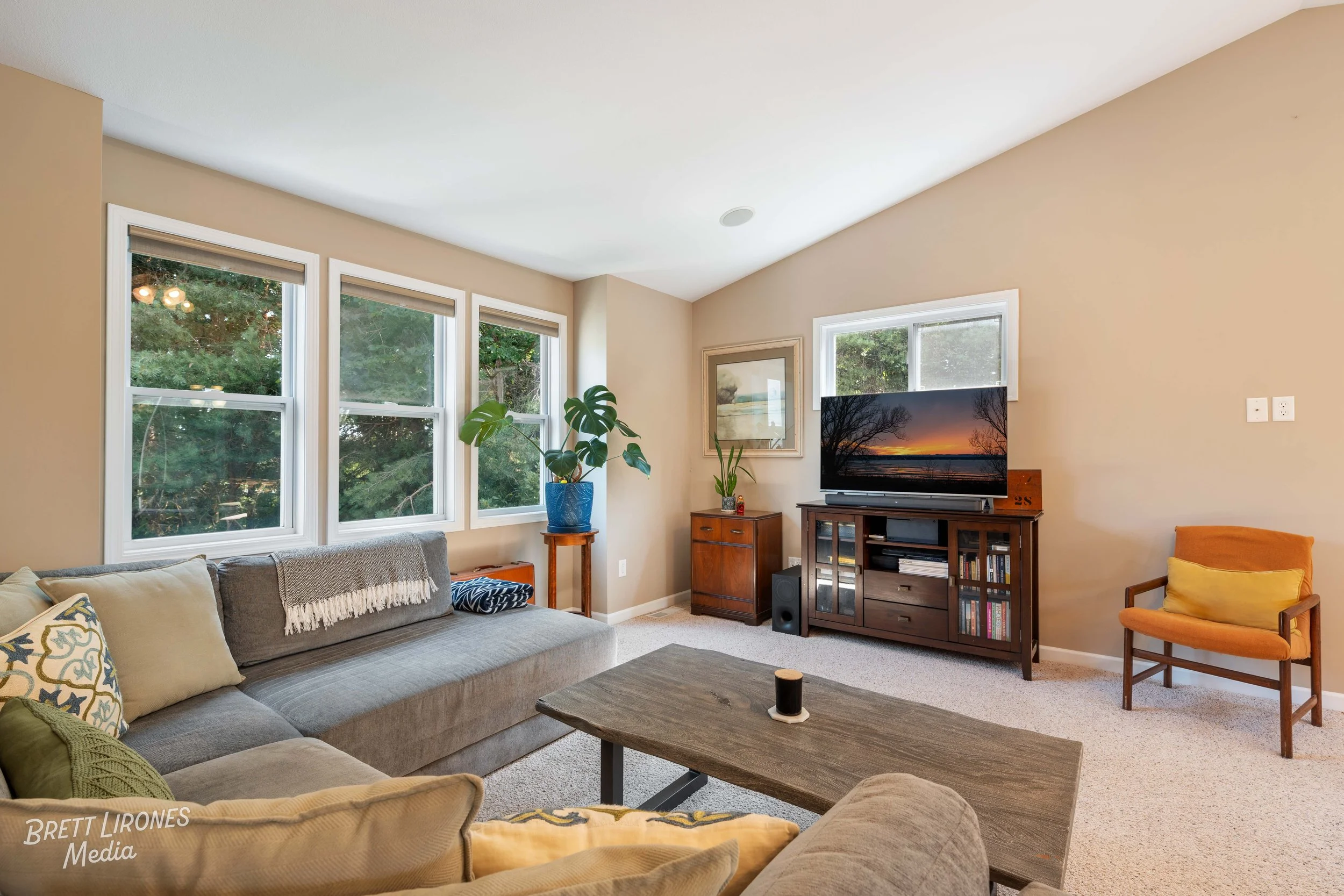 Living room with beige walls, large windows, a gray sectional sofa with pillows, a wooden coffee table, a TV on a wooden stand, a small cabinet, a potted plant, and a single orange armchair.