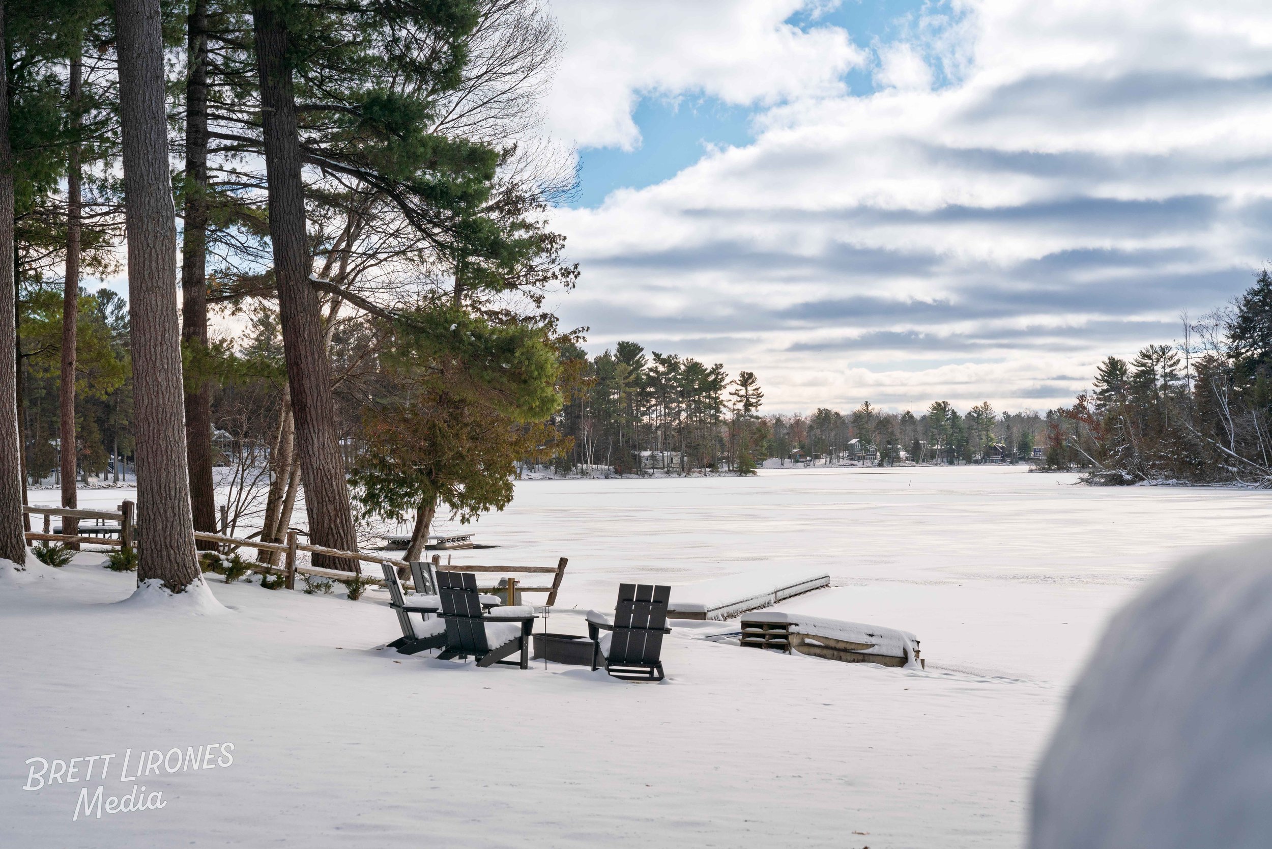 A snowy lakeside scene with trees and Adirondack chairs on the snow, a wooden dock extending onto the frozen lake, and a cloudy sky.