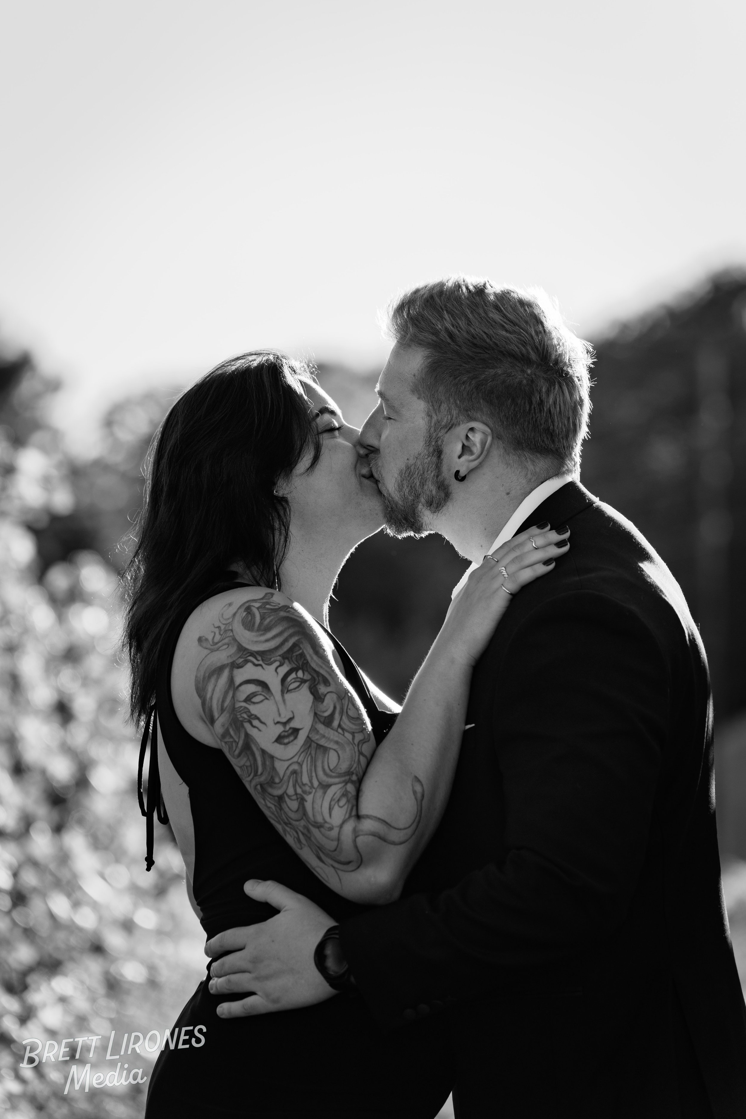 A black-and-white photo of a couple kissing outdoors, the woman with dark hair and a large tattoo on her arm, the man with short hair and a beard, both dressed formally.