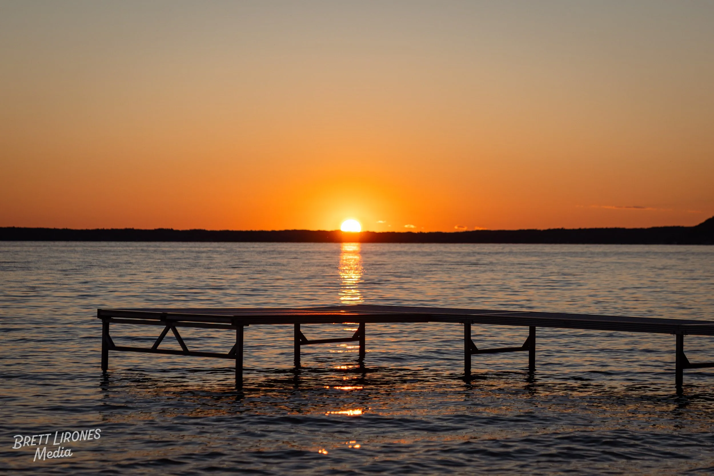 Sunset over a large body of water with a wooden dock partially submerged in the water in the foreground. The sky is clear with warm orange and yellow hues.