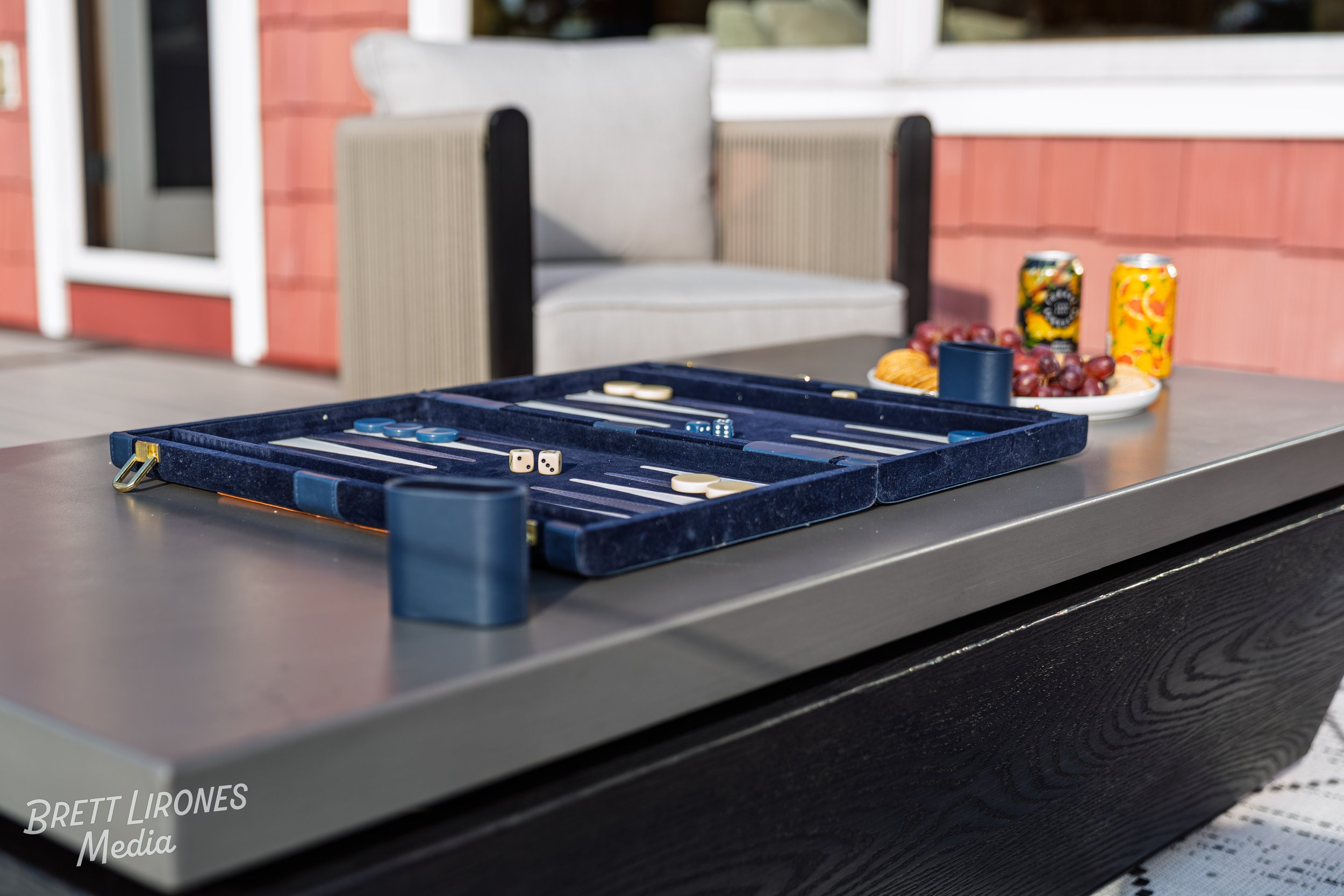 A backgammon game set up on an outdoor table, with a plate of grapes and snacks, and two cans of fruit-flavored drinks in the background.